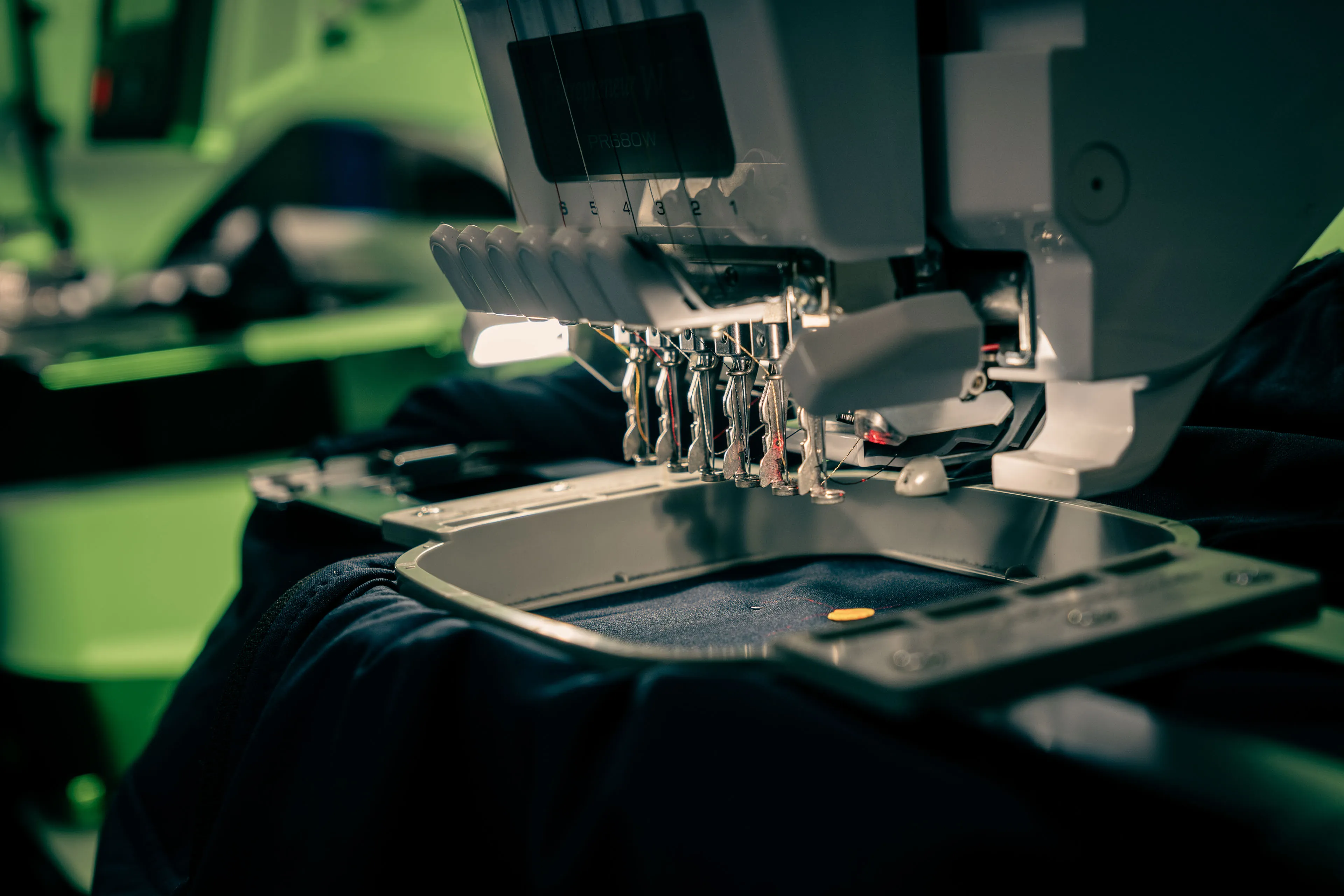 Close-up of a modern embroidery machine with multiple needles poised over dark fabric in an industrial setting, highlighting precision and technology.