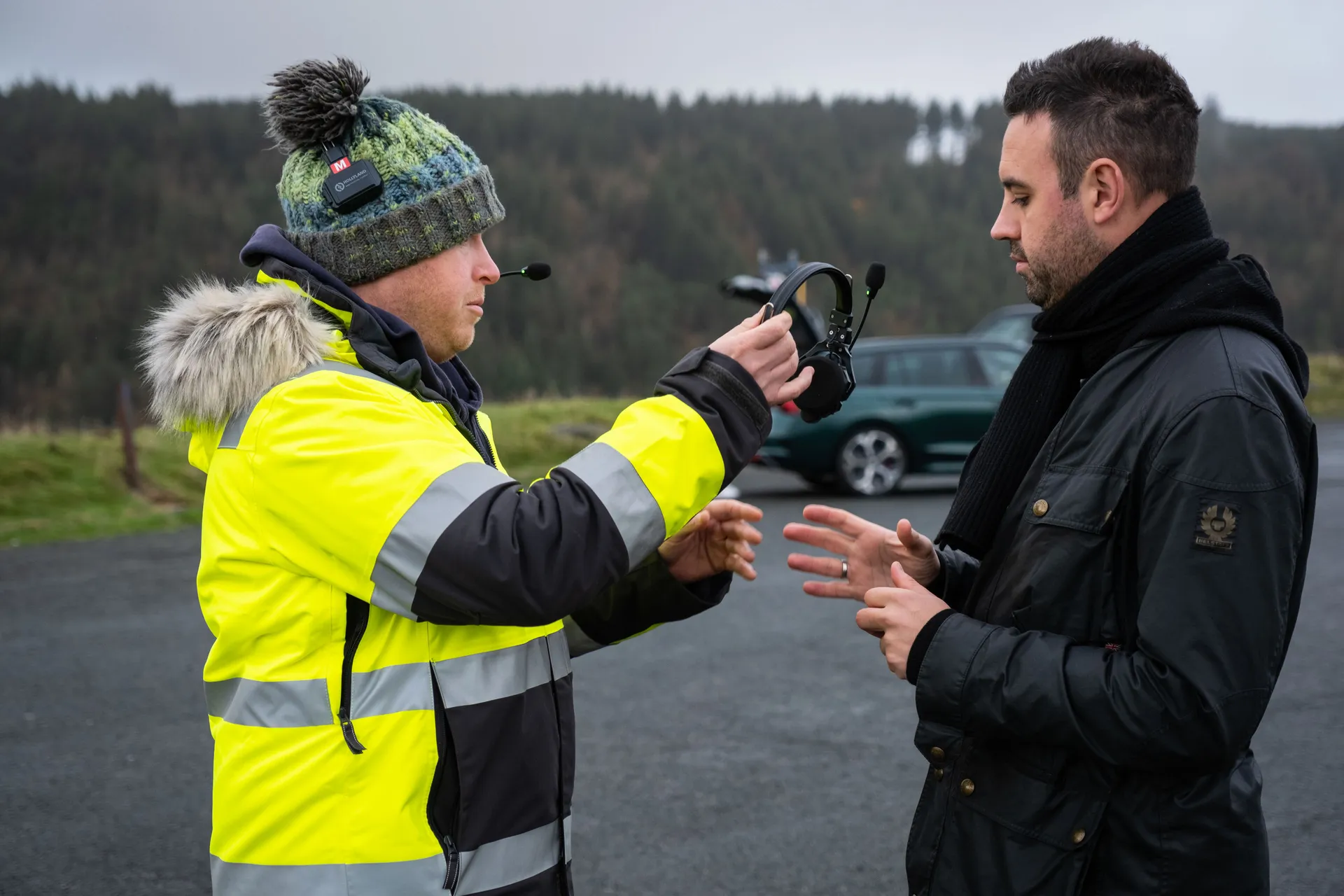 Two men in a conversation on a cloudy day; one in a high-visibility jacket and beanie holds headphones, while the other wears a dark coat, with a green car and forest in the background.