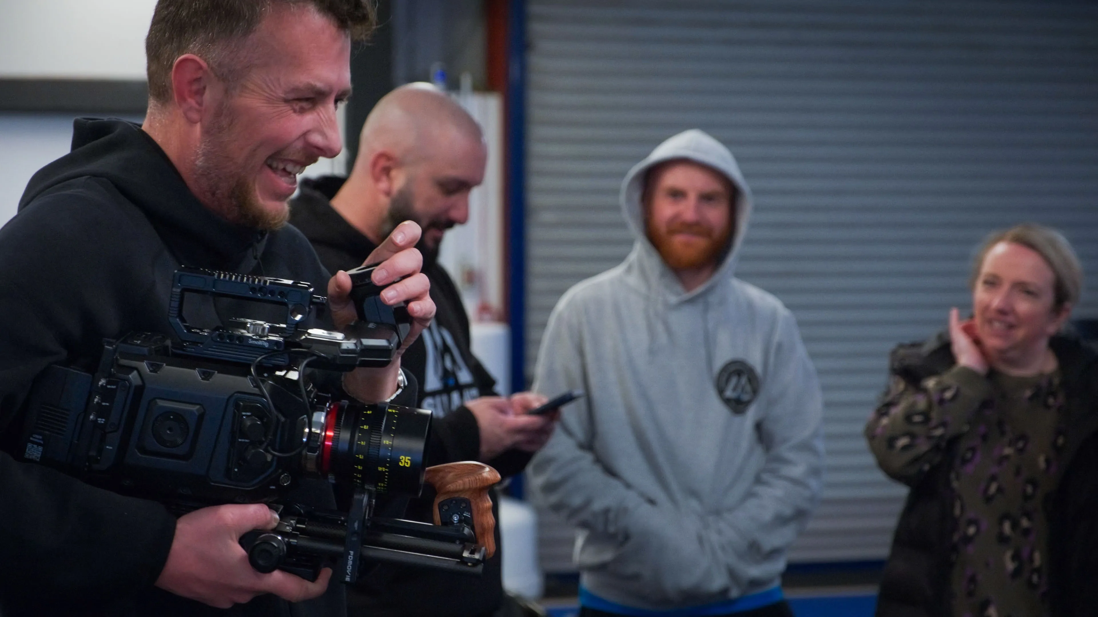 A man holding a video camera laughs during filming, surrounded by three casually dressed people. The scene conveys a relaxed and cheerful atmosphere.
