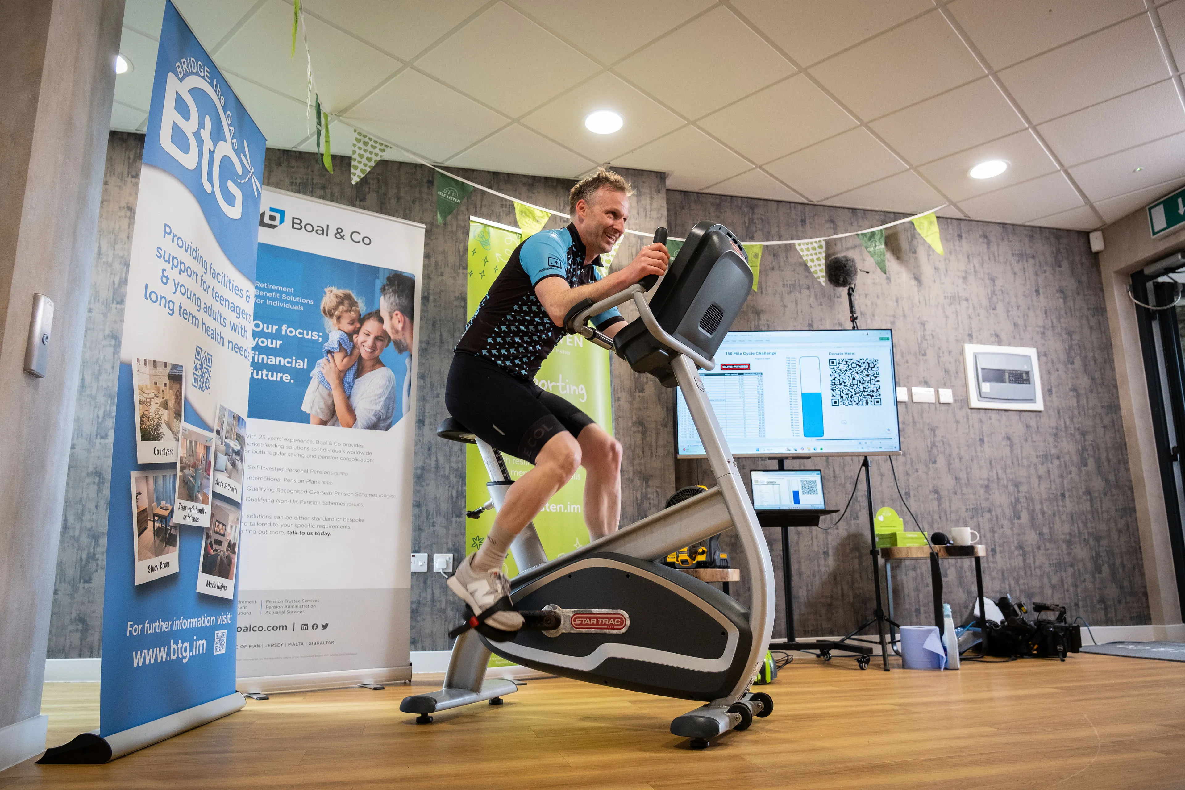 John Beaty from DotPerformance cycling on a static bike in DotPerformance cycling kit at the Boal & Co charity event on the Isle of Man, with a fundraising tracker visible on a screen in the background
