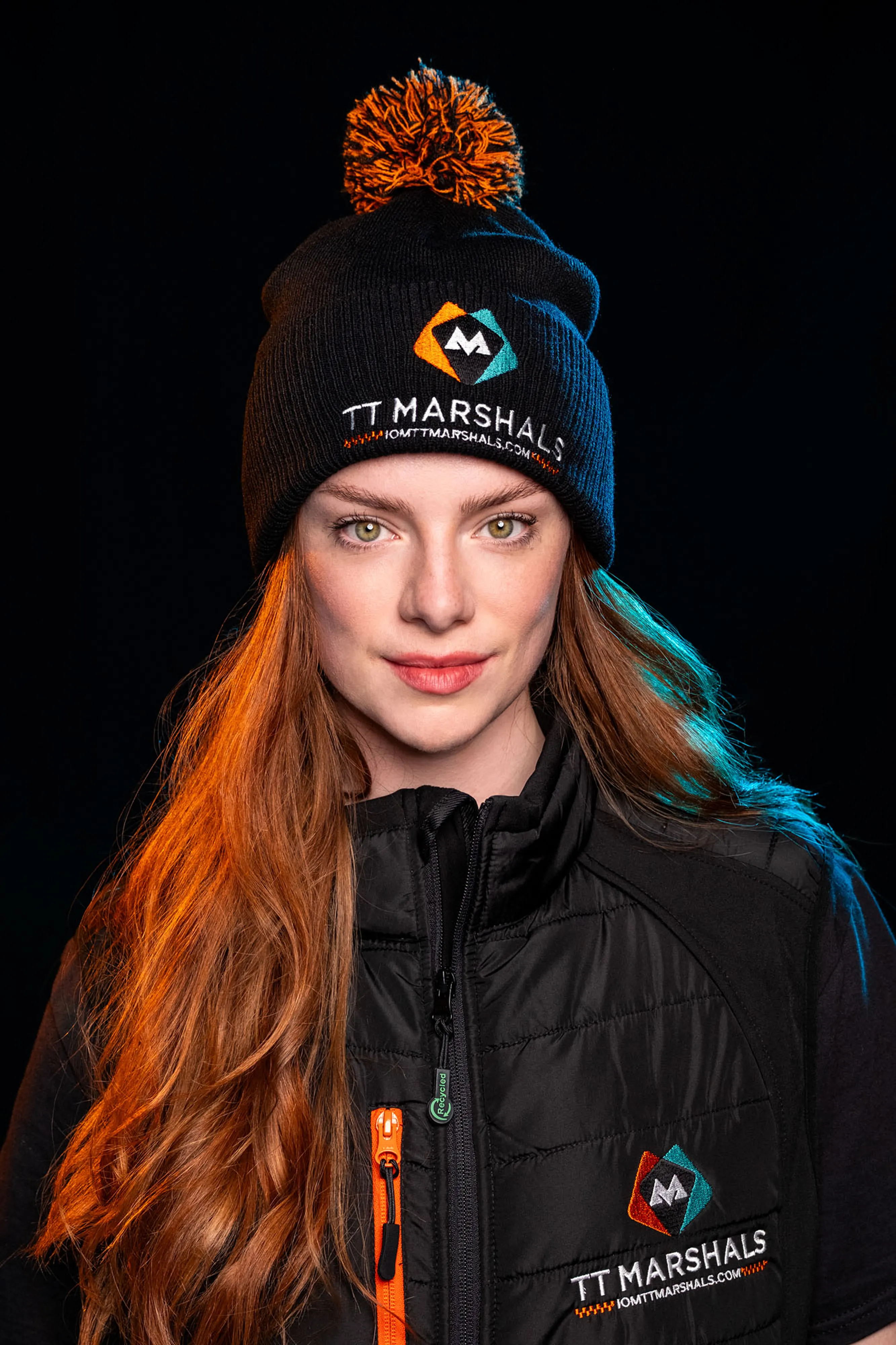 Ella Garvey wearing the official TT Marshals 2026 black bobble hat and softshell padded gilet, featuring the TT Marshals logo. Exclusive marshal merchandise for the Isle of Man TT 2026.
