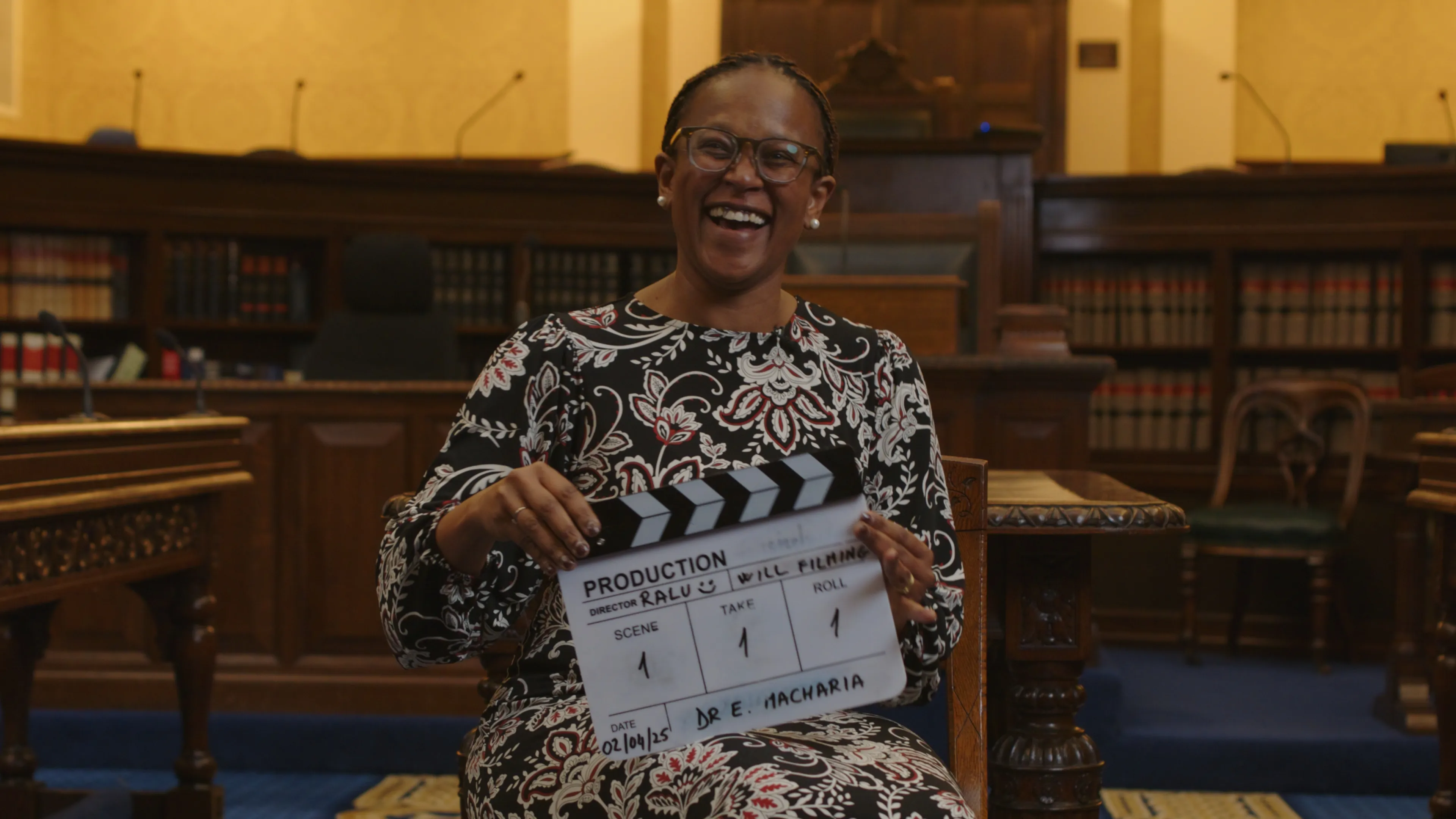 Smiling woman in a patterned dress holds a clapperboard in a library-themed room. The atmosphere is warm and welcoming, conveying joy and professionalism.