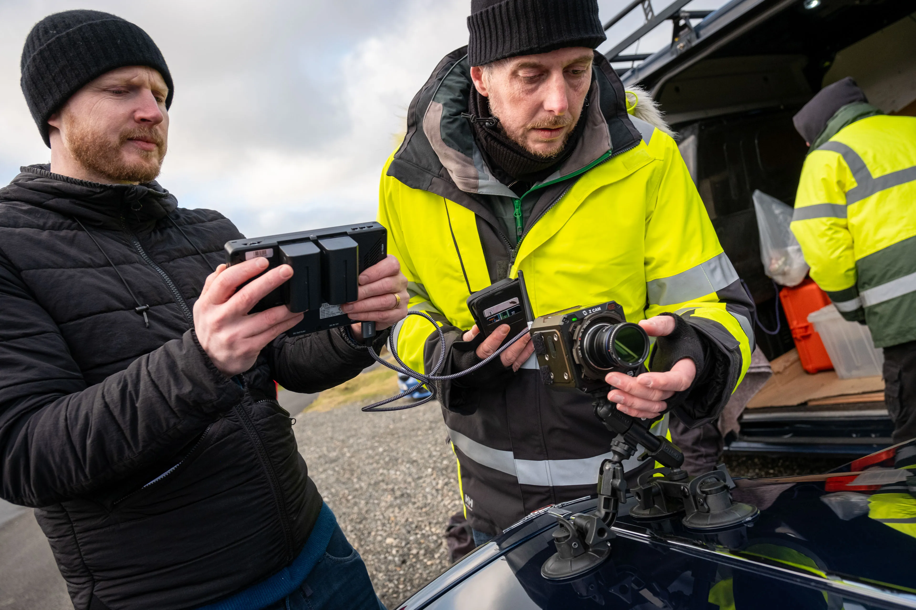 Two men in winter clothing work together. One holds a camera, checking its settings, while the other operates a monitor. They're focused and collaborative.