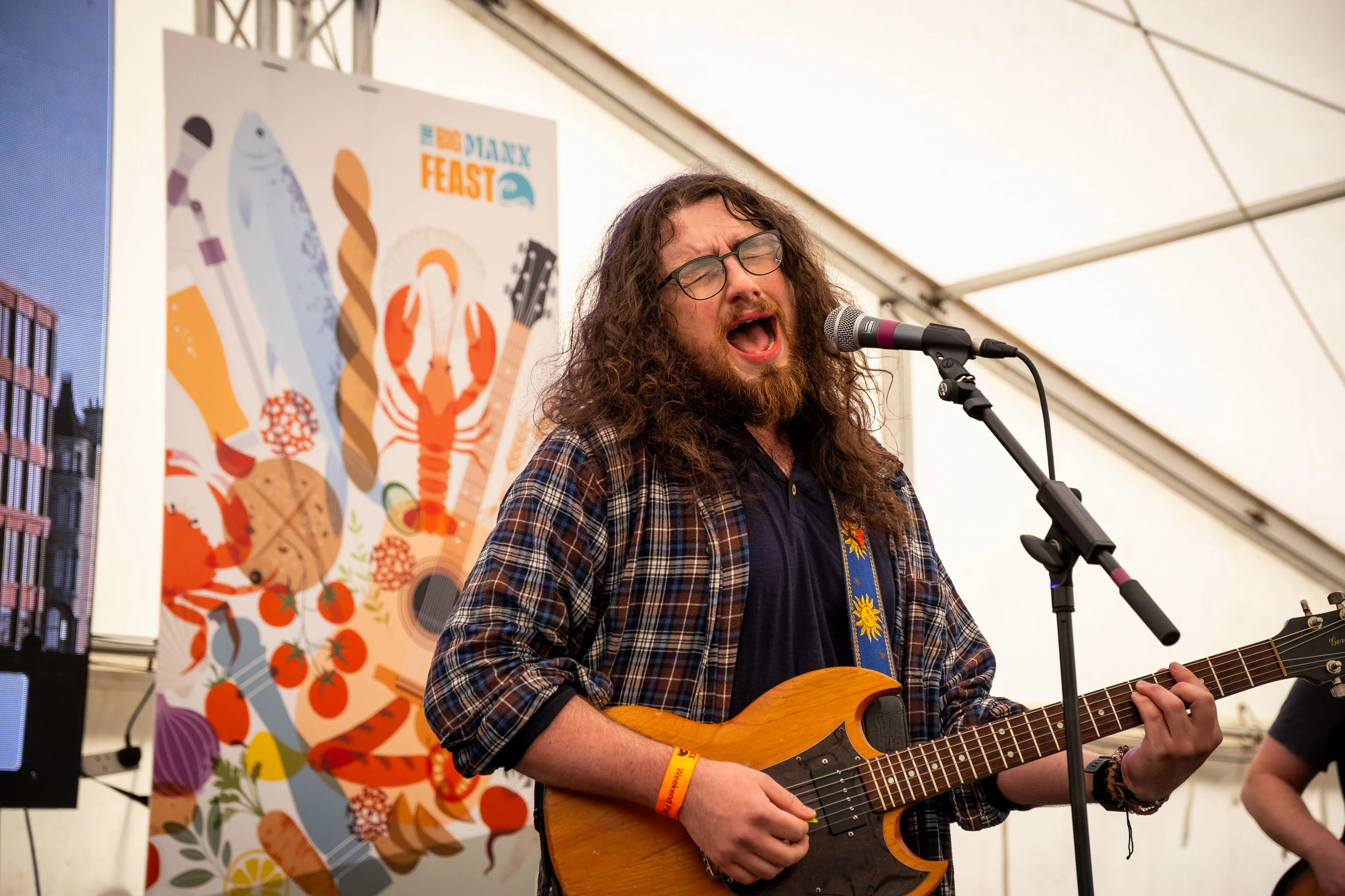 Harry Roe playing guitar and singing on stage in front of the Big Manx Feast banner
