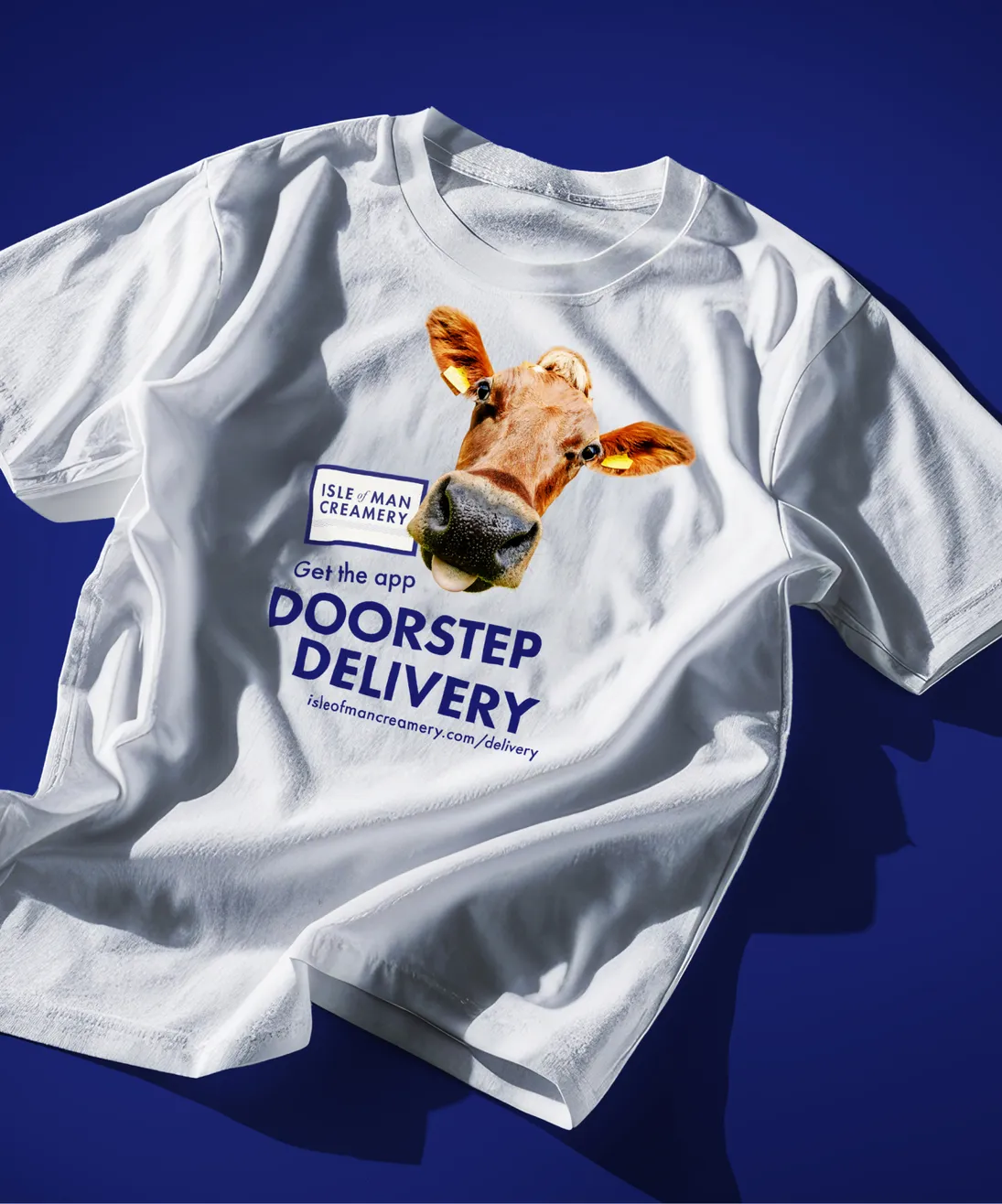 A white t-shirt laid flat on a blue background, printed with a close-up photo of a Jersey cow's face, the Isle of Man Creamery logo, and bold blue text reading "Get the app – Doorstep Delivery" with the URL isleofmancreamery.com/delivery