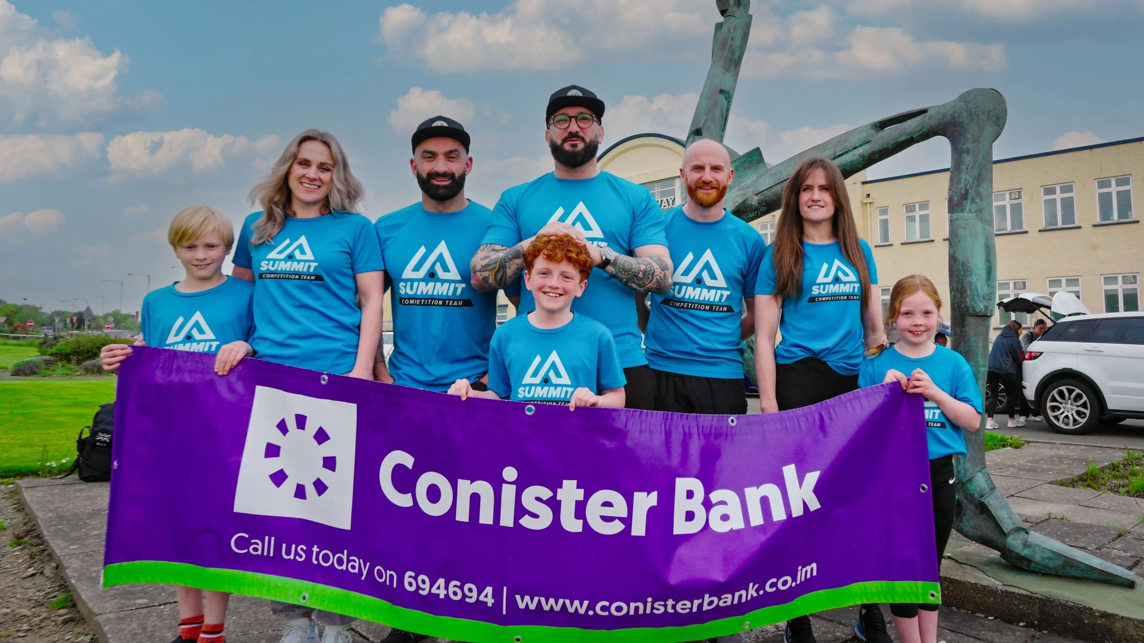 A diverse group of eight people wearing blue "Summit" shirts stand outdoors, smiling and holding a purple "Conister Bank" banner. A large sculpture is in the background.
