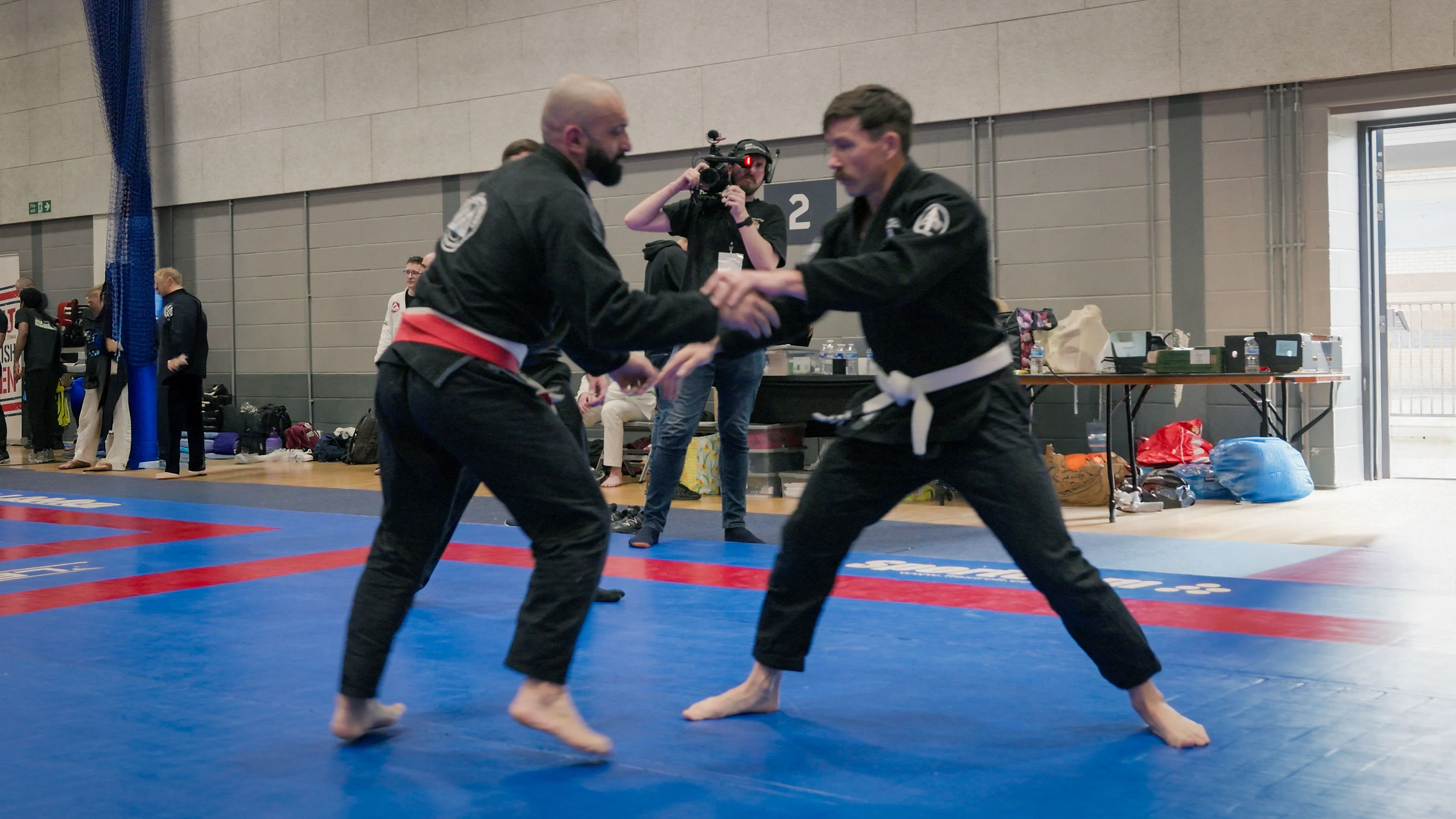 Two men in black martial arts gi, one with a red belt and the other with a white belt, grapple on a blue mat in a gym while a photographer captures the scene.