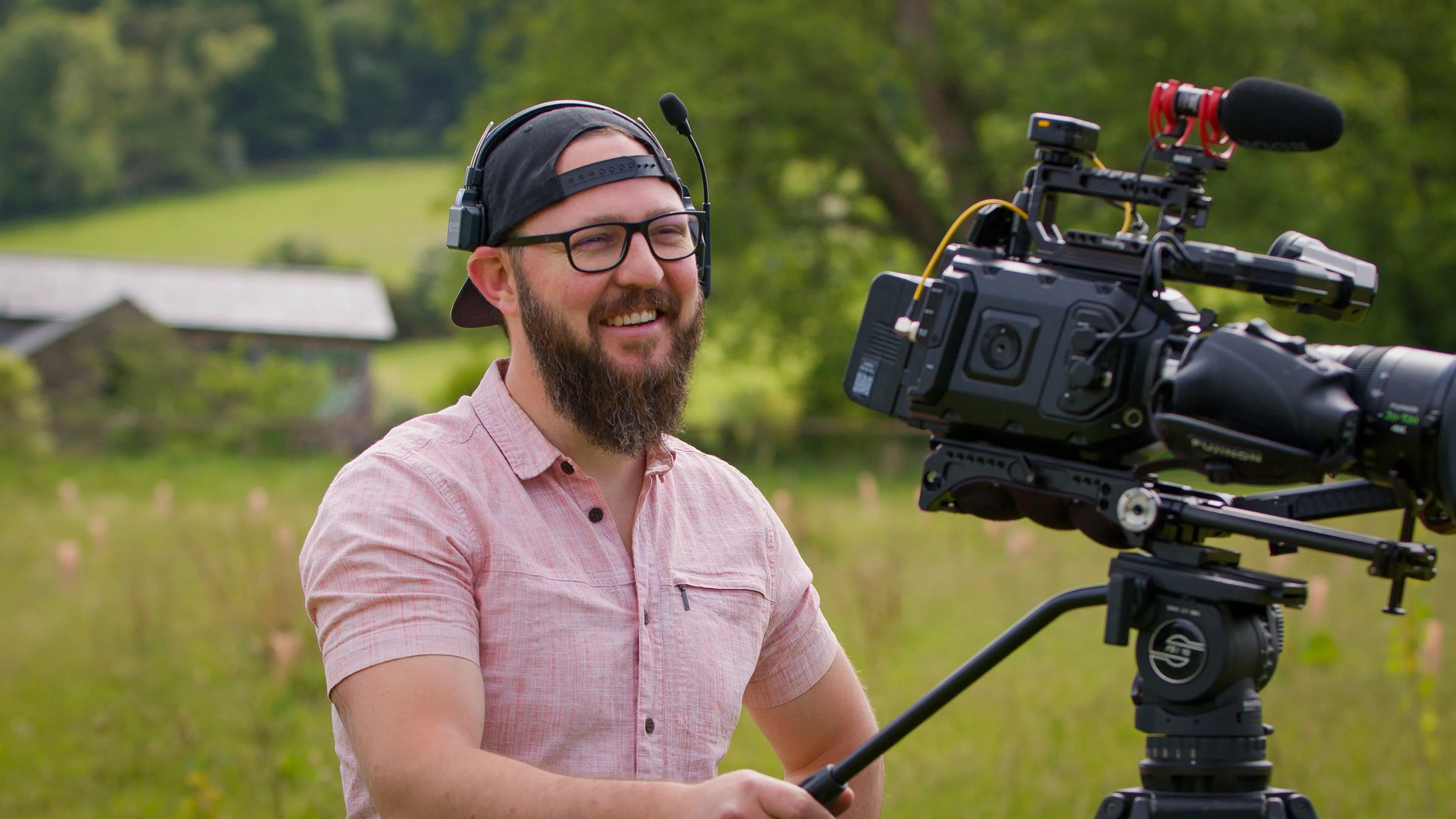 A bearded man with glasses and a backward cap smiling while operating a professional video camera outdoors in a green field, conveying a relaxed and joyful mood.