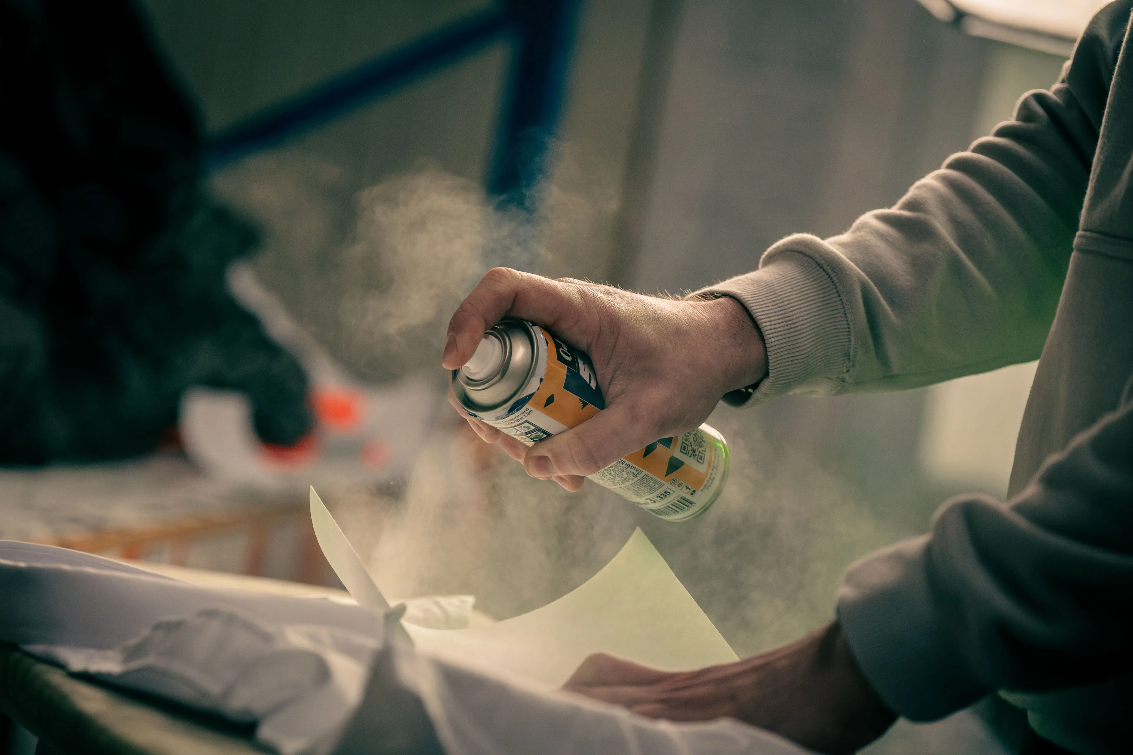 A hand sprays adhesive from an aerosol can onto a large sheet of paper. The mist creates a soft, focused effect. The mood is concentrated and artistic.