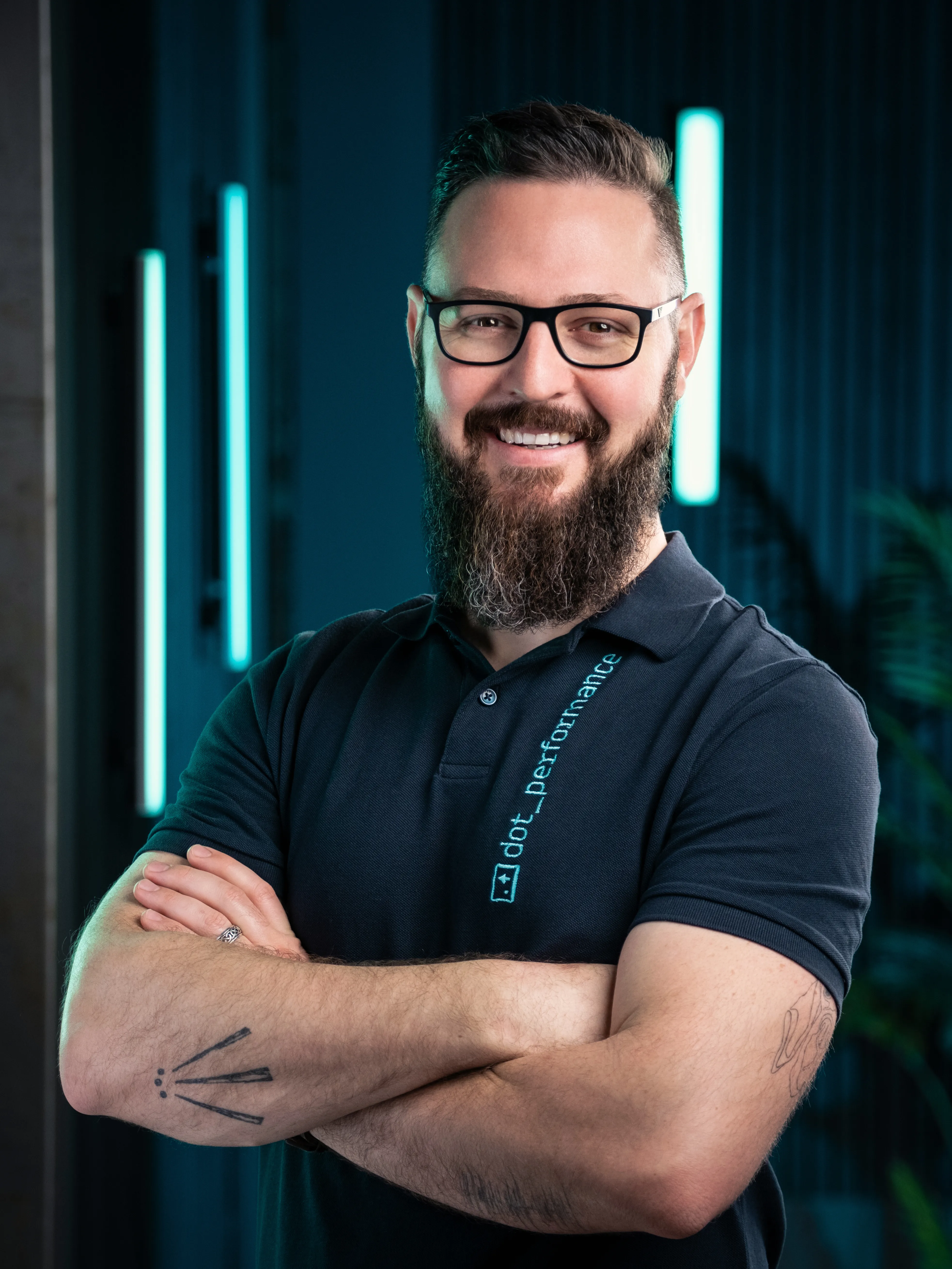 Nico, photographer and cinematographer at Dot Performance, smiling with crossed arms as he poses for headshot wearing branded polo shirt, against dark background with blue neon lights.