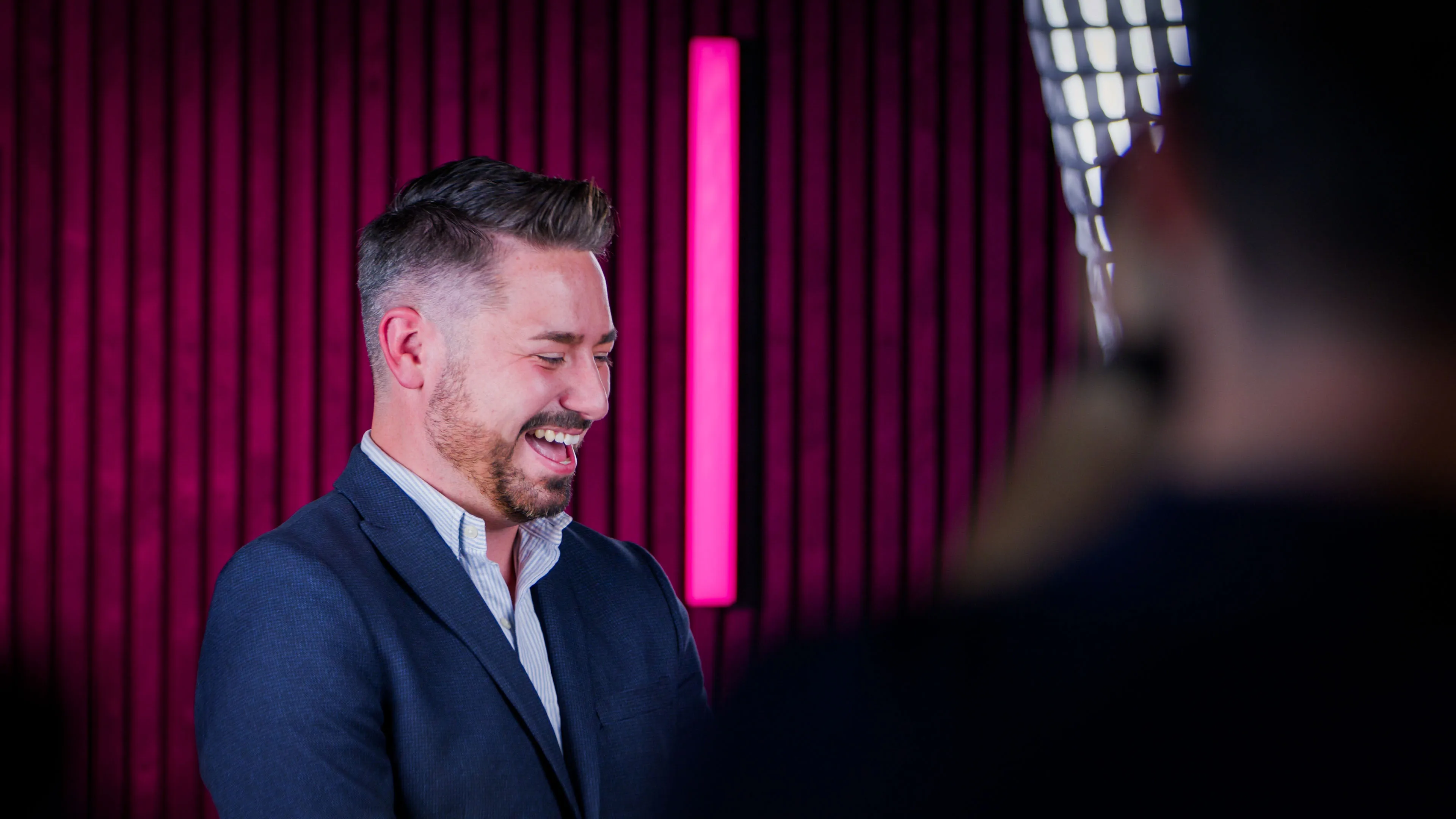 Man in a blue blazer and white shirt smiles warmly against a modern setting with vertical magenta lights and dark striped walls, conveying a cheerful mood.