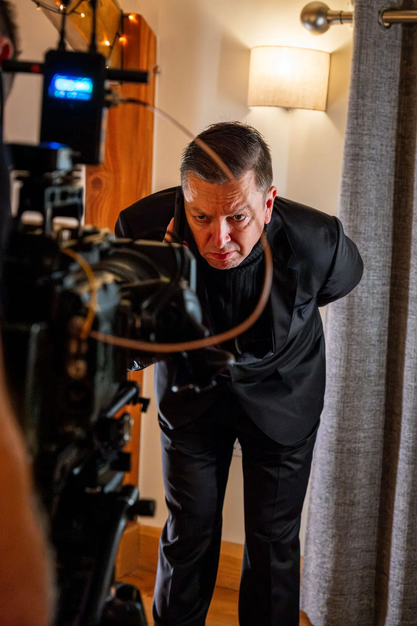 Man in a suit, leaning towards a camera with a serious expression. Warm indoor lighting, curtain and lamp in the background, creating a focused atmosphere.