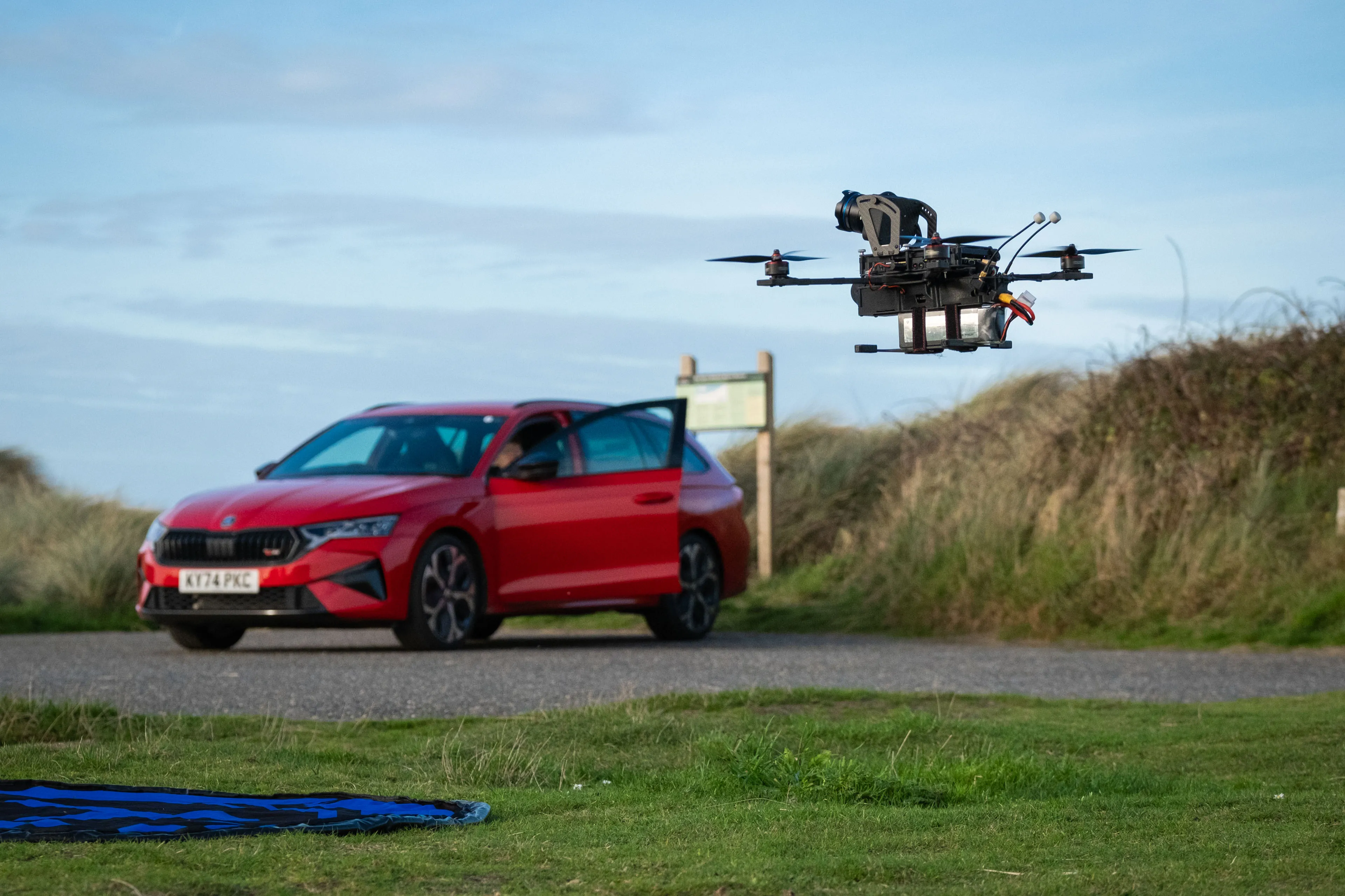 A drone hovers low over grassy terrain, with a red car in the background. The car's door is open.