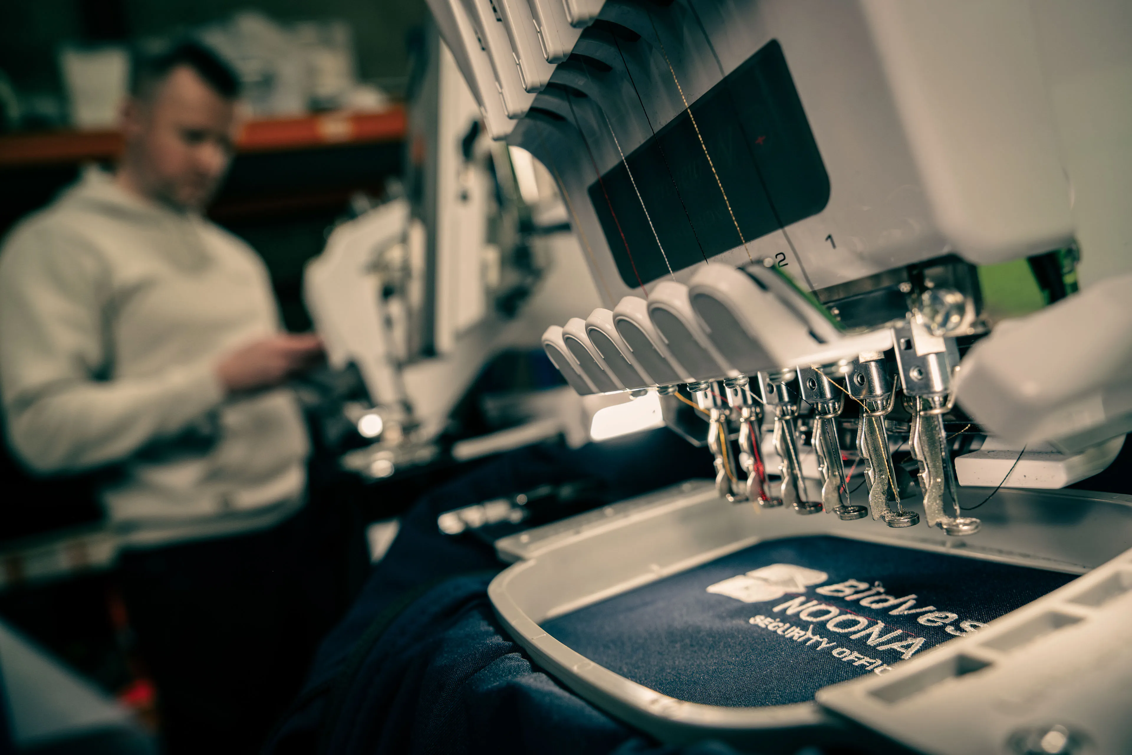 Close-up of an embroidery machine stitching a logo onto fabric, with a blurred person in the background. The scene conveys precision and focus.