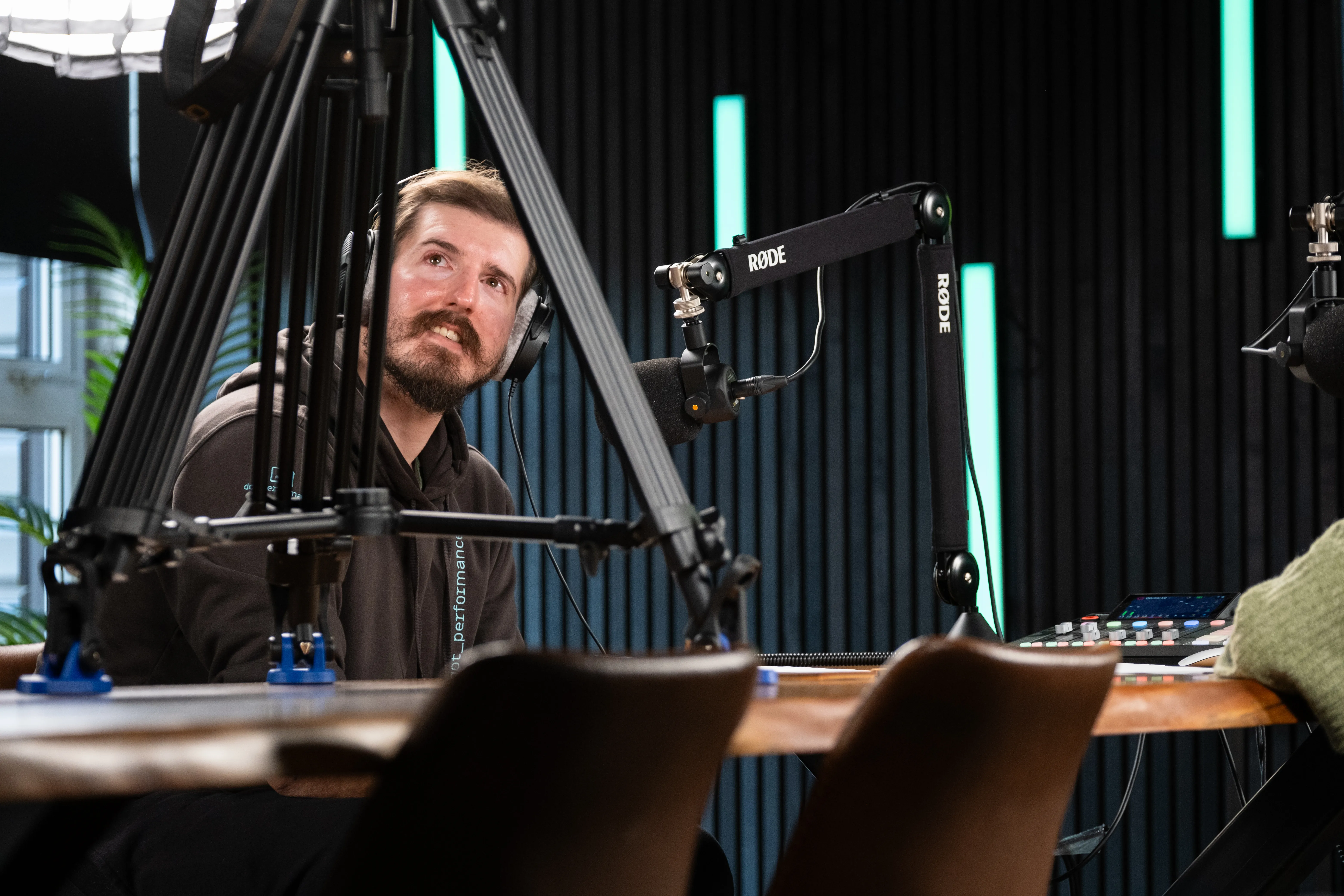 Will Oates wearing headphones at the recording desk in DotPerformance studios on the Isle of Man, surrounded by Røde microphones, camera rigs, and audio mixing equipment