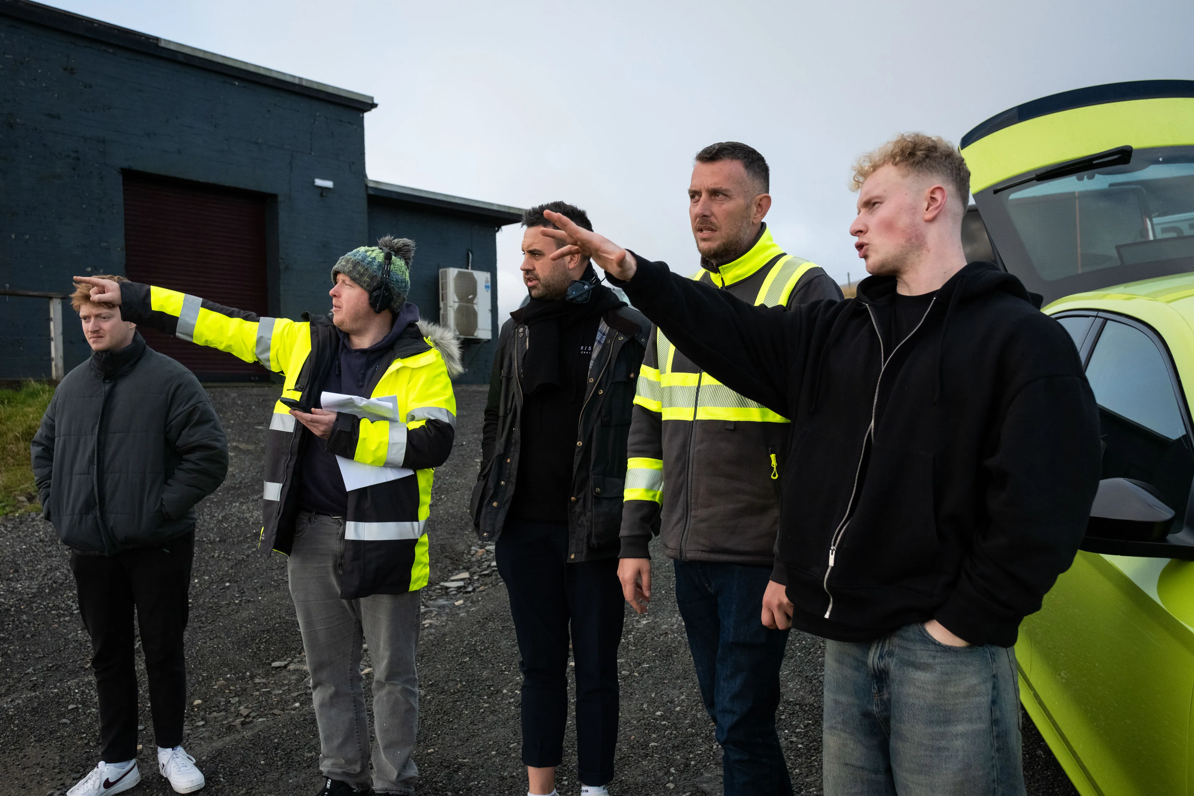 Five men stand near a building and a neon green vehicle, pointing and looking intently in one direction, wearing casual and high-visibility jackets.