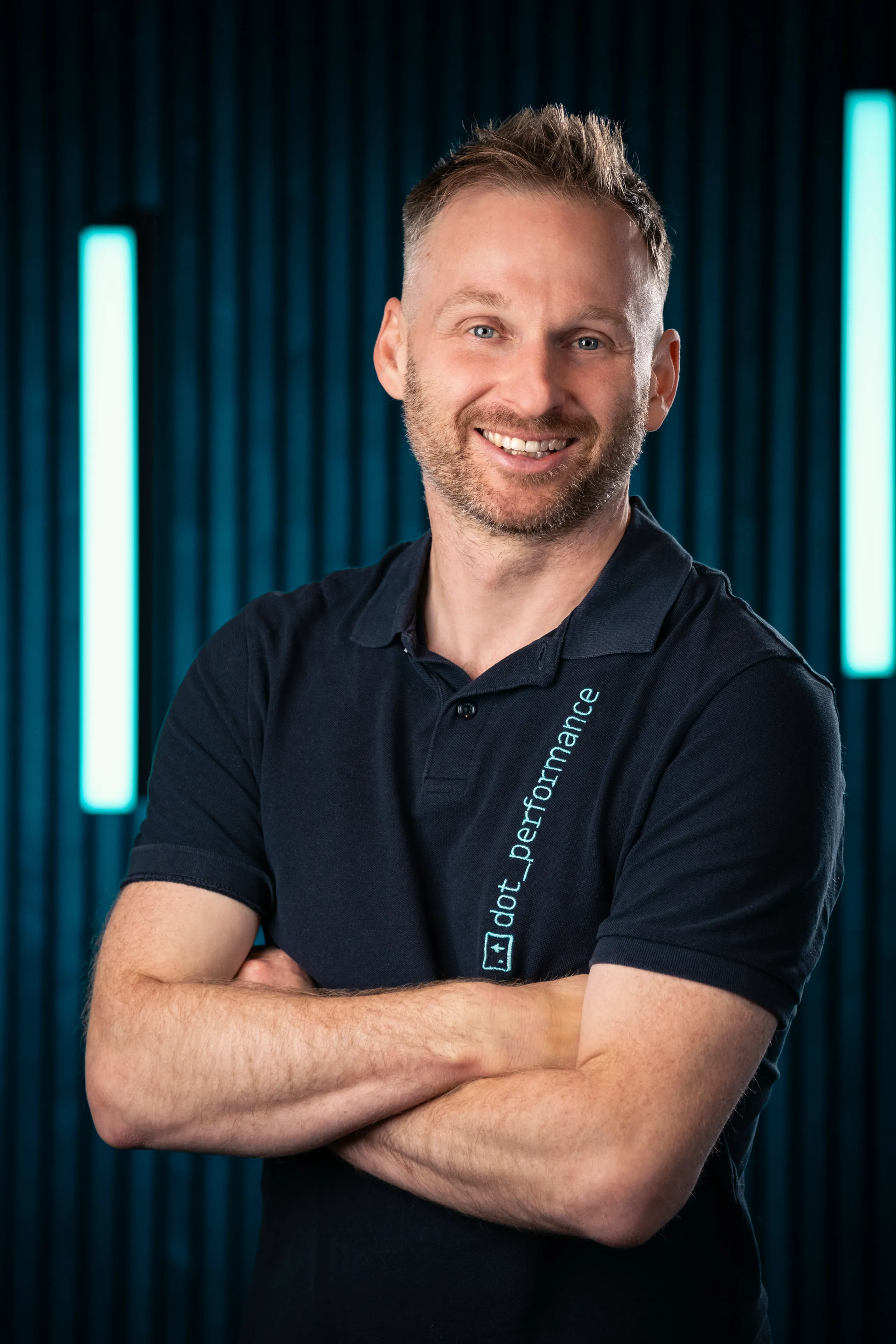 John Beaty, Head of Systems at DotPerformance, smiling with arms crossed wearing a DotPerformance branded polo shirt at DotPerformance studios on the Isle of Man, against a dark background with teal neon lighting