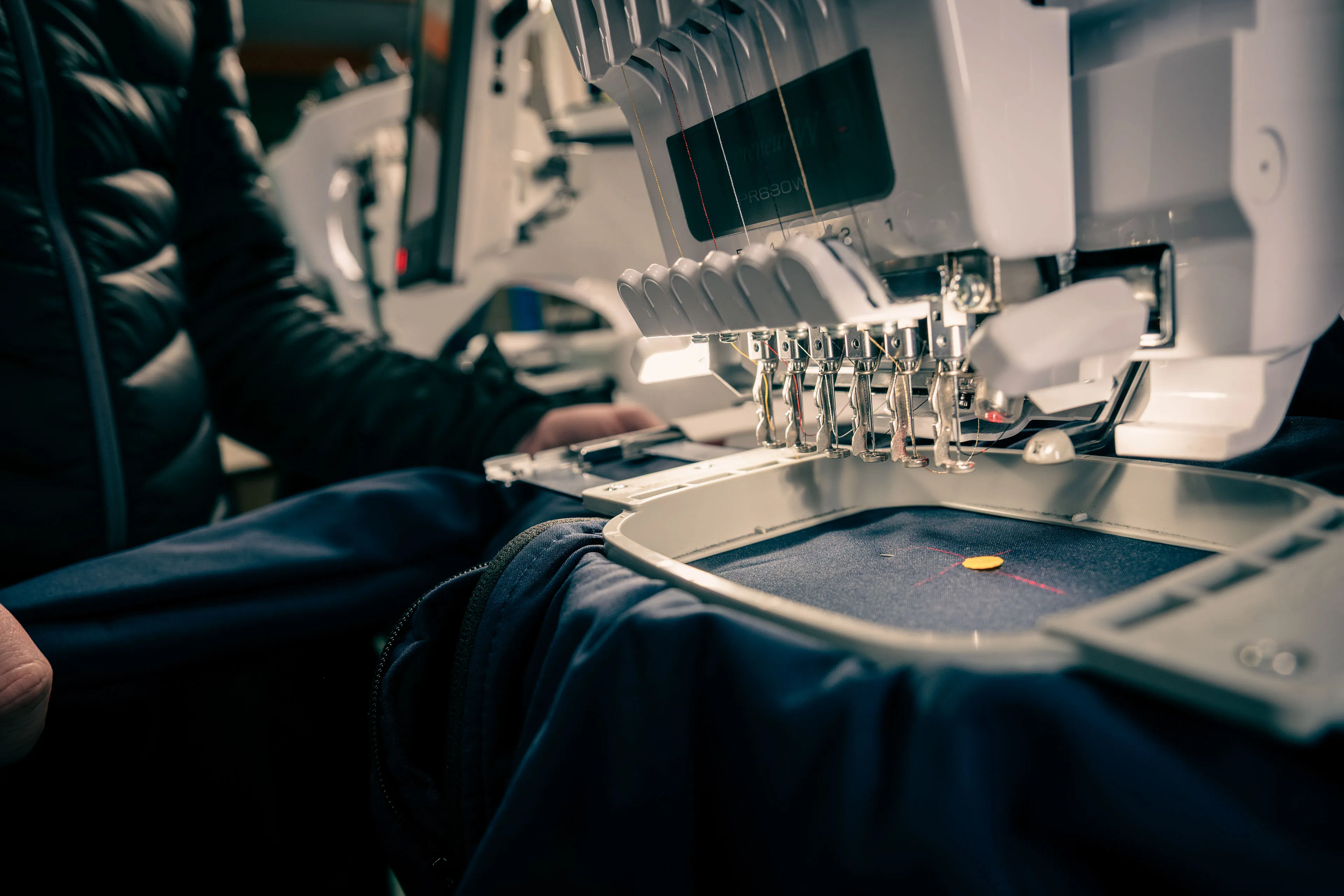 A close-up of an embroidery machine working on dark fabric. Needles are threading, with a person in a jacket adjusting the material, creating a focused and industrious atmosphere.