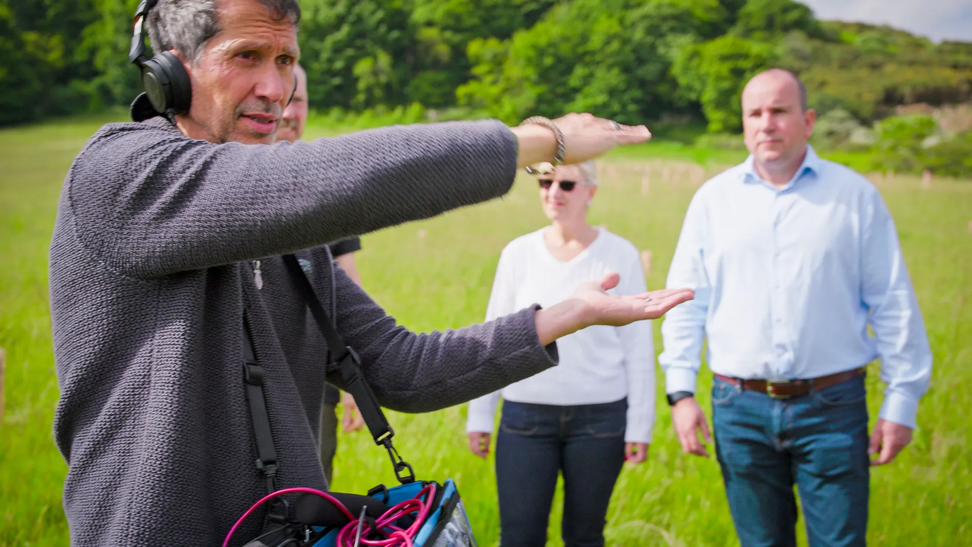 A man wearing headphones gestures with both hands while directing two people standing in a green field. The scene conveys focus and collaboration.