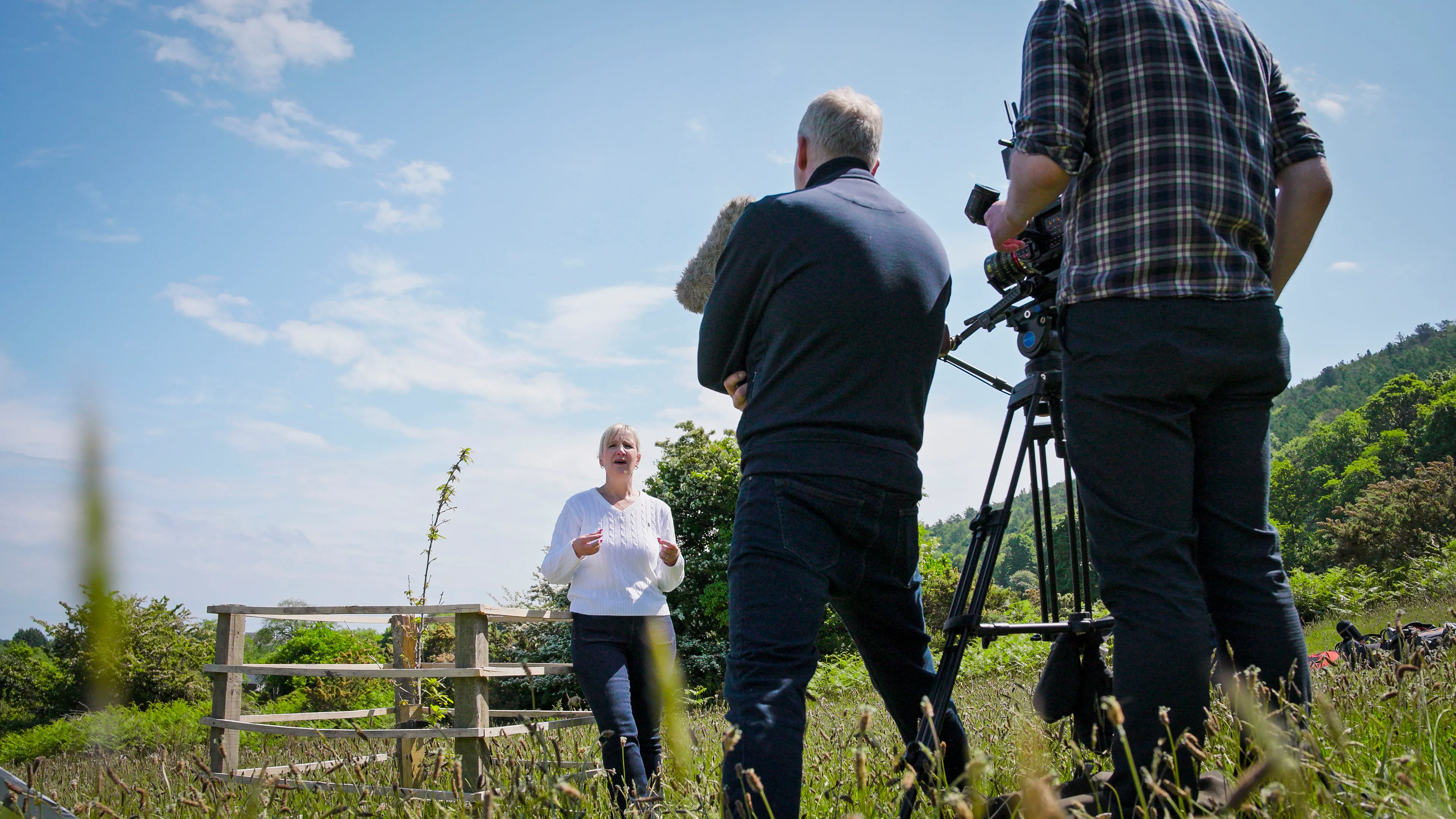 A woman speaks energetically outdoors with a small wooden fence behind her. Two men film her with a camera and microphone, set against a blue sky and green hills.