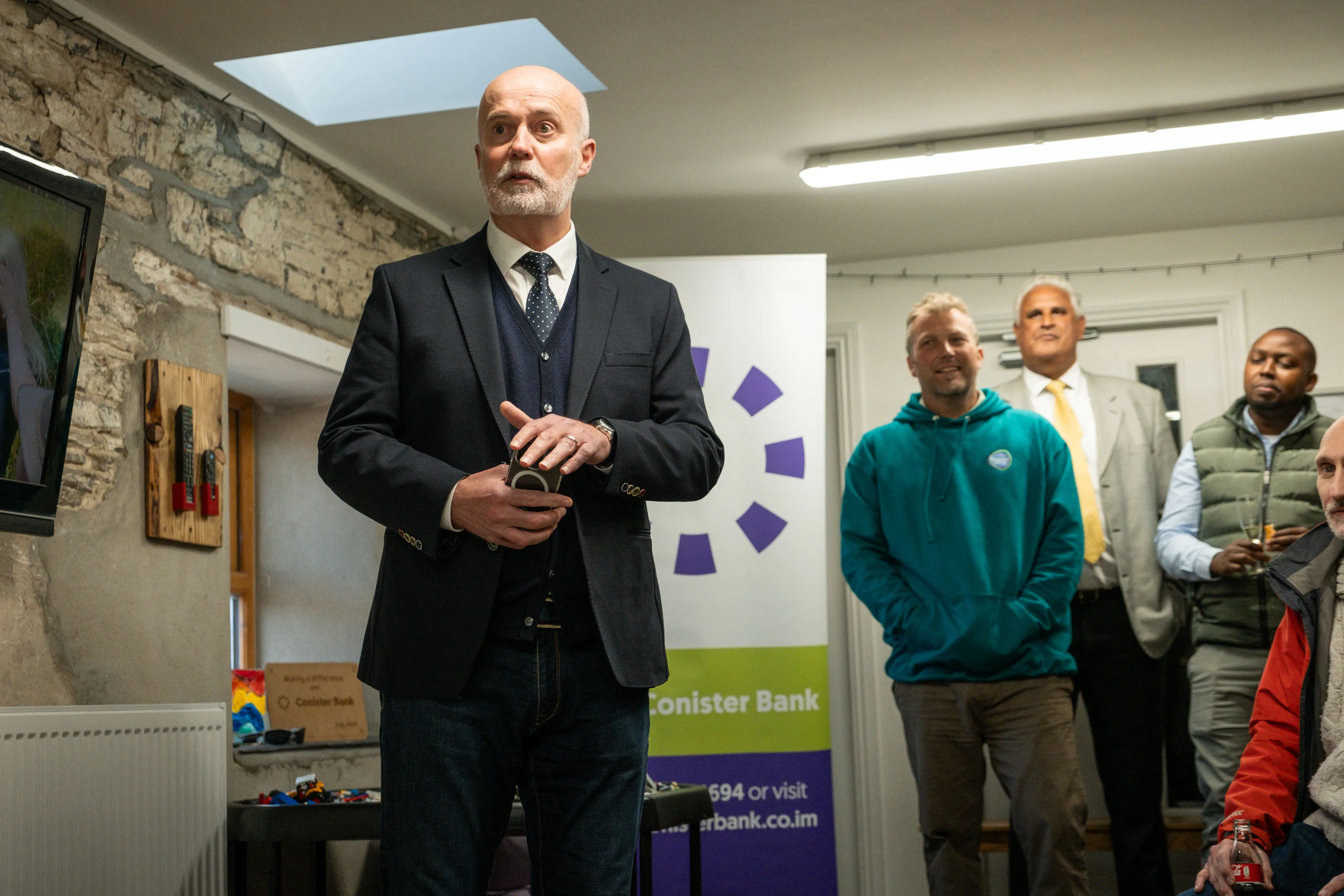 Douglas Grant addressing staff and guests at an indoor event at The Children's Centre, Isle of Man, with a Conister Bank banner visible in the background, filmed by DotPerformance