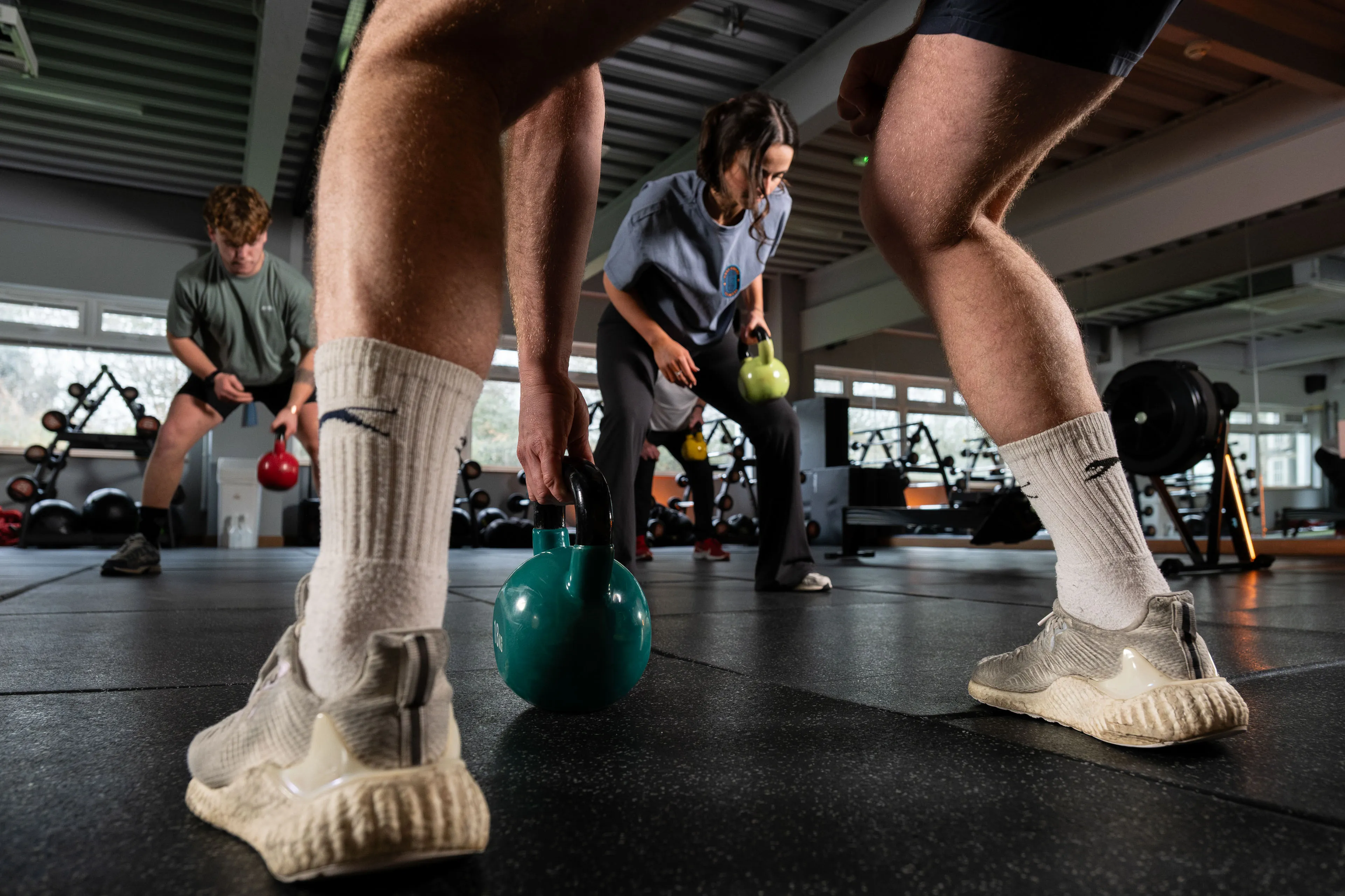Three people in a gym focus on kettlebell exercises. A close-up of legs in athletic gear is in the foreground, conveying energy and concentration.