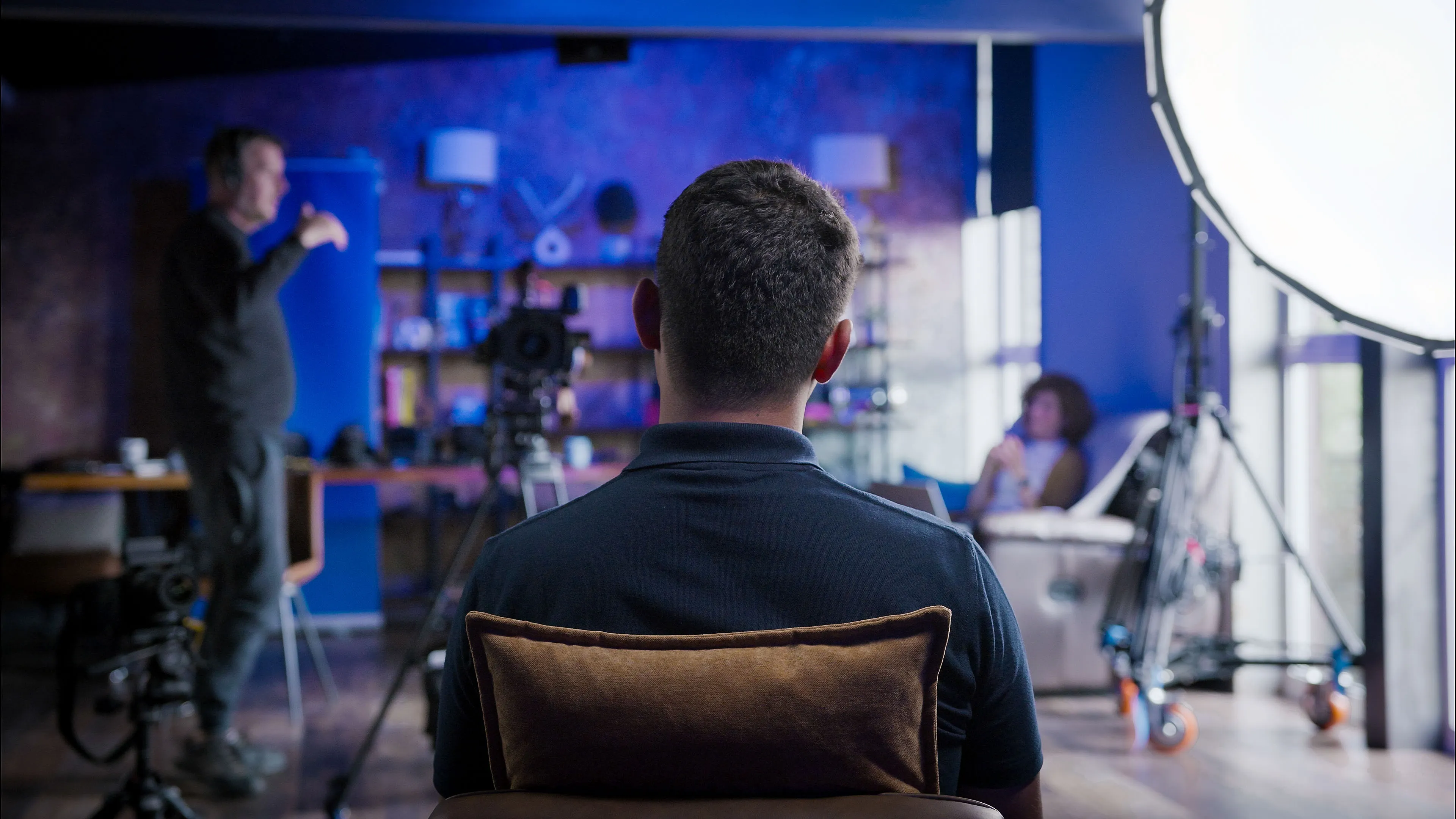 A man sits with his back to the camera in a filming studio, facing bright lights and equipment. Another person sits on a chair, creating a focused, professional atmosphere.