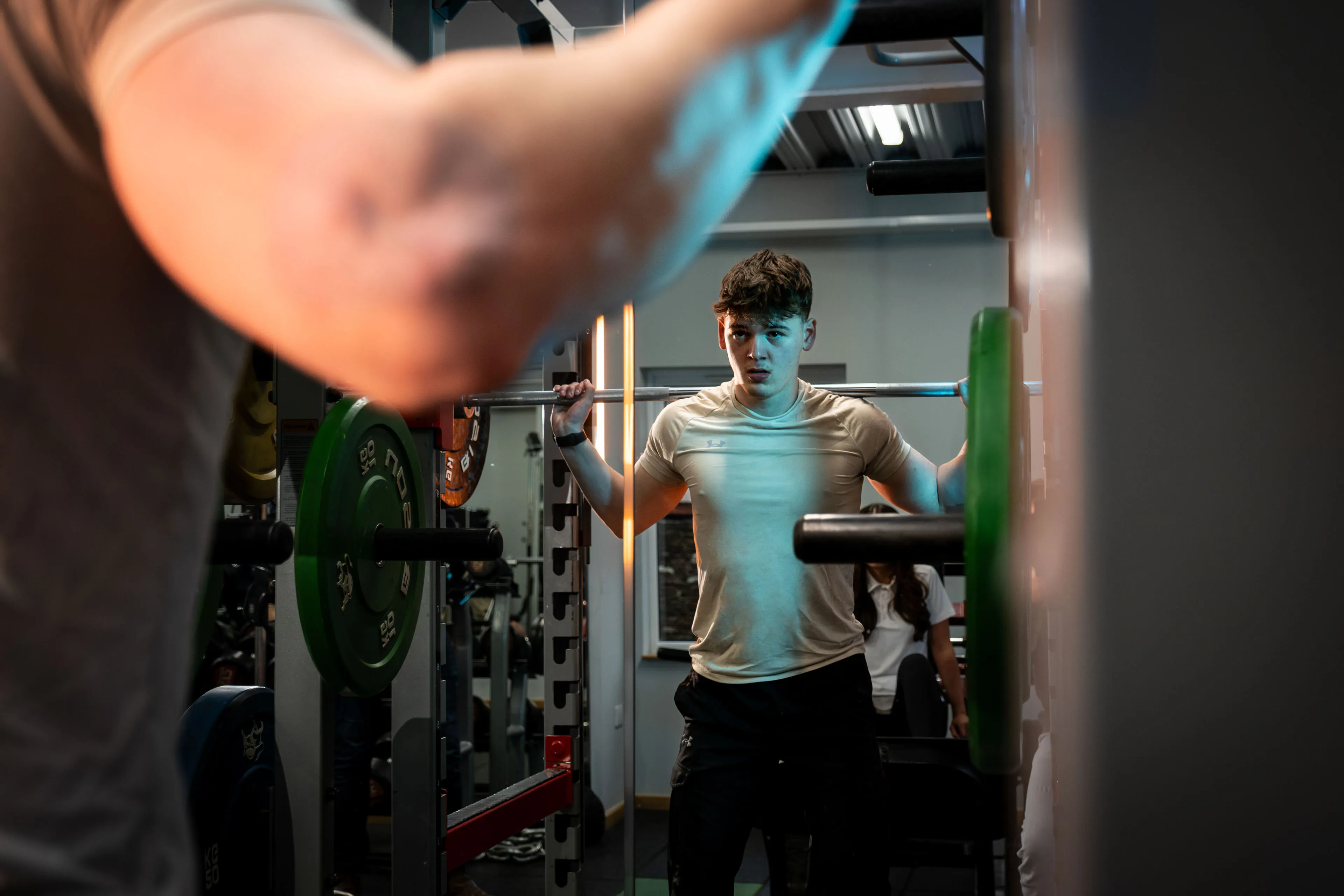 Young man focused on lifting weights in a dimly lit gym. He stands at a squat rack, eyes forward, with determination and intensity.
