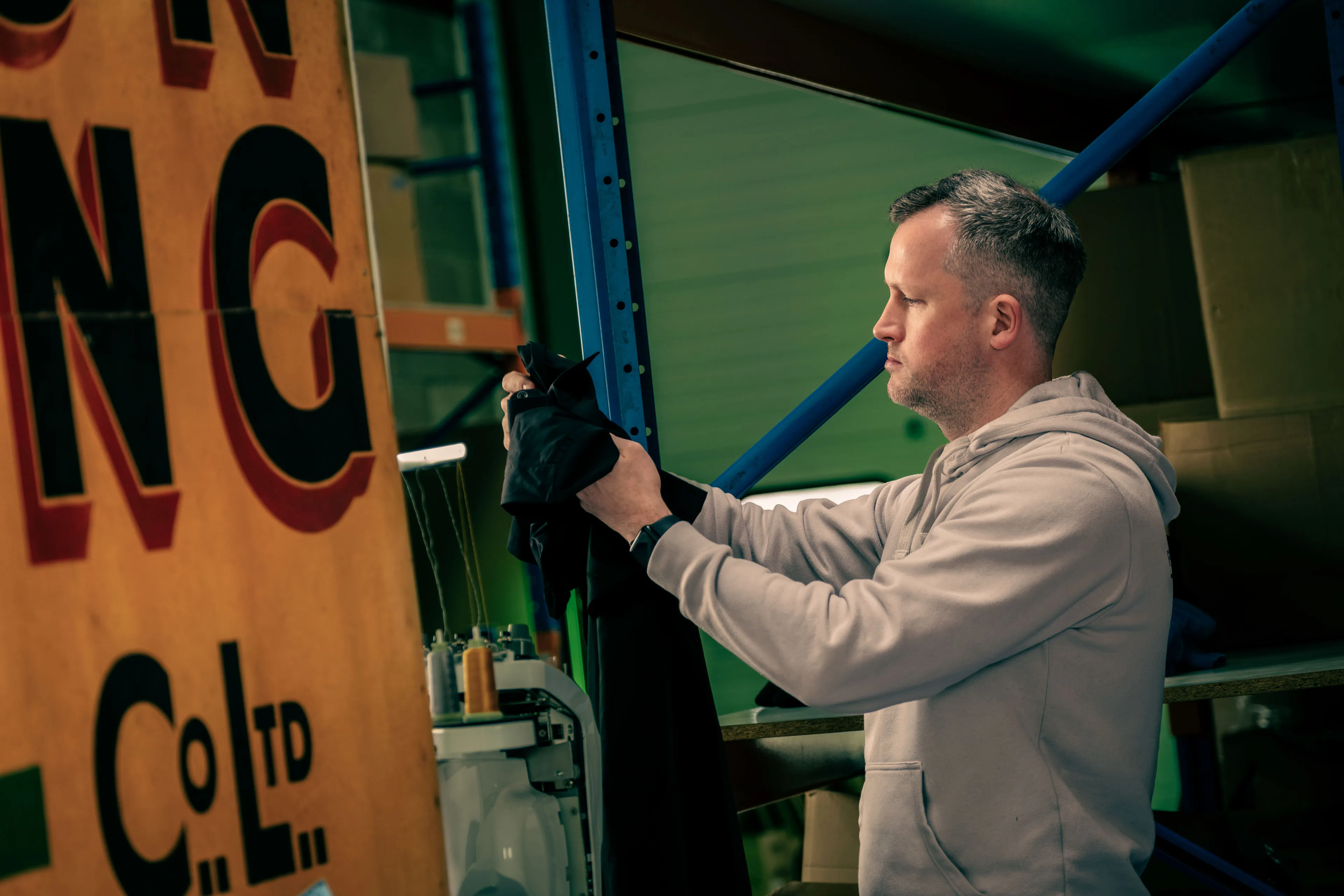A man wearing a hoodie examines a black garment in a warehouse. He stands near a sewing machine and a large vintage sign, surrounded by shelves and boxes.