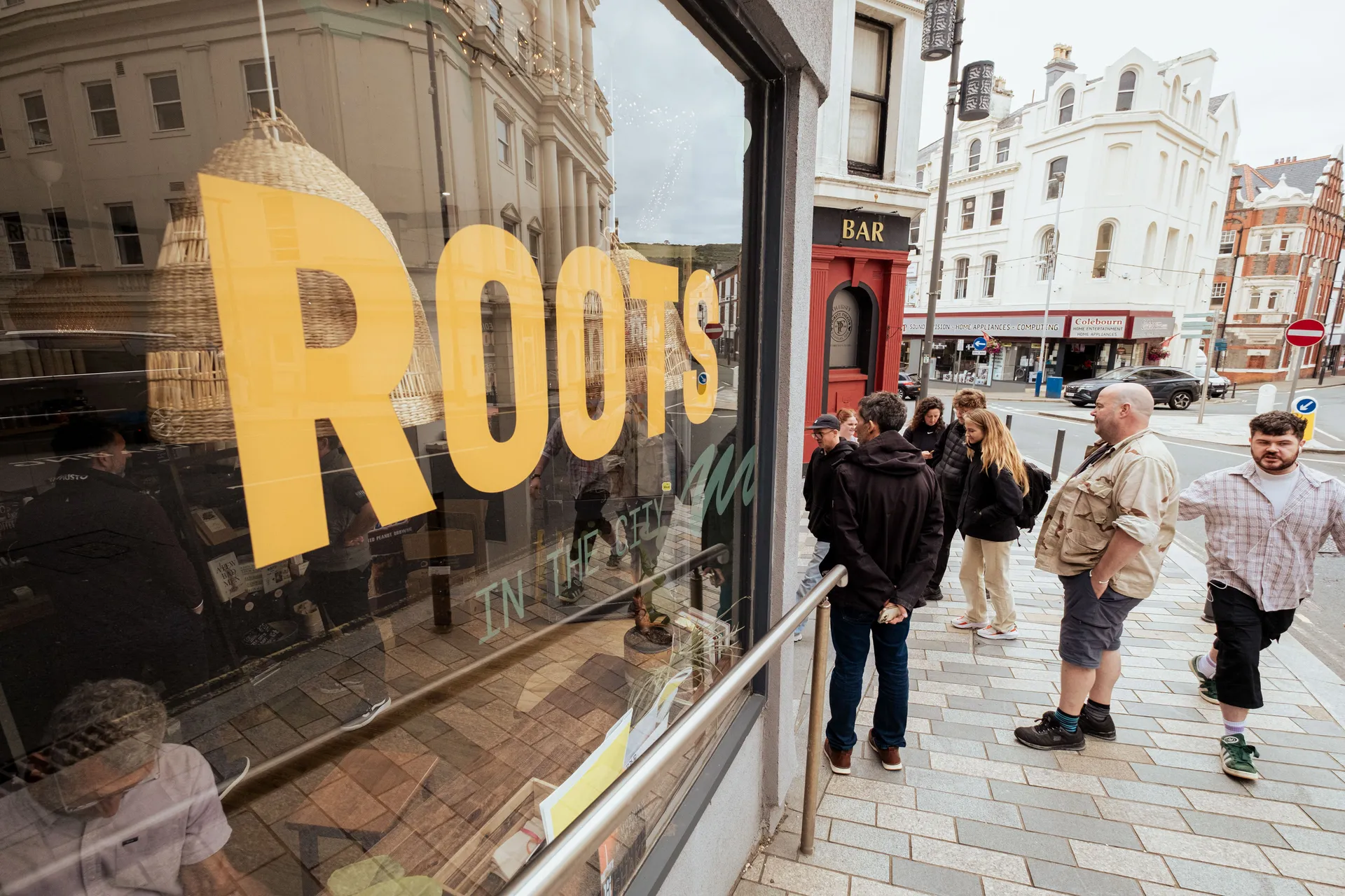 People stand outside a coffee shop with a large window sign reading "ROOTS." The street is busy with pedestrians, creating a lively urban scene.