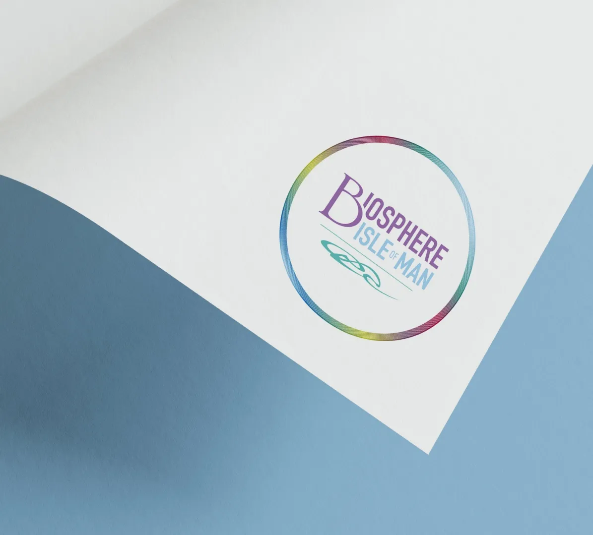 Biosphere Isle of Man logo printed on a curved white card showing the circular rainbow ring mark with Celtic motif and purple and teal lettering, designed by DotPerformance