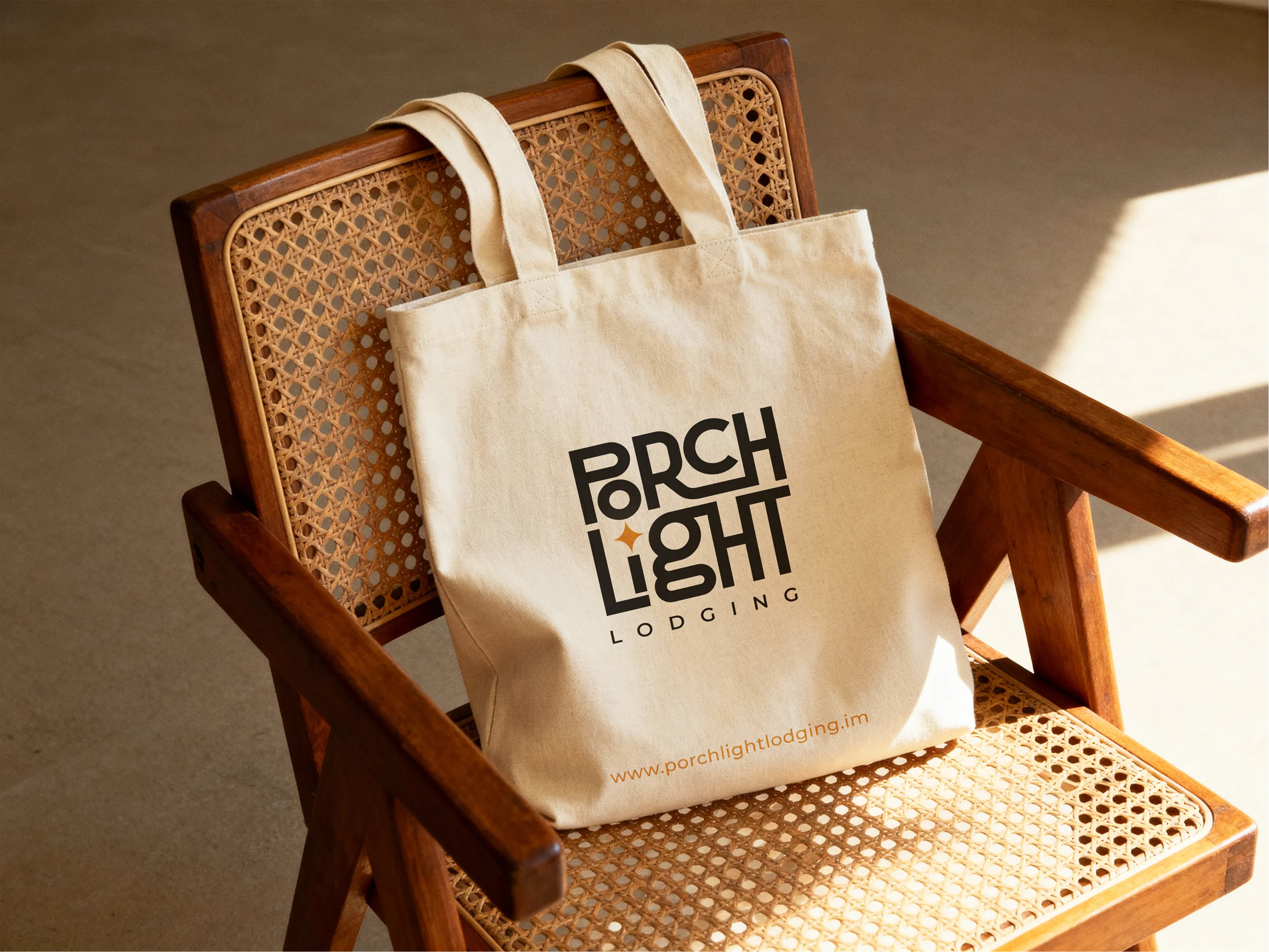 Cream canvas tote bag printed with the PorchLight Lodging logo, resting on a wooden cane-back chair in warm sunlight