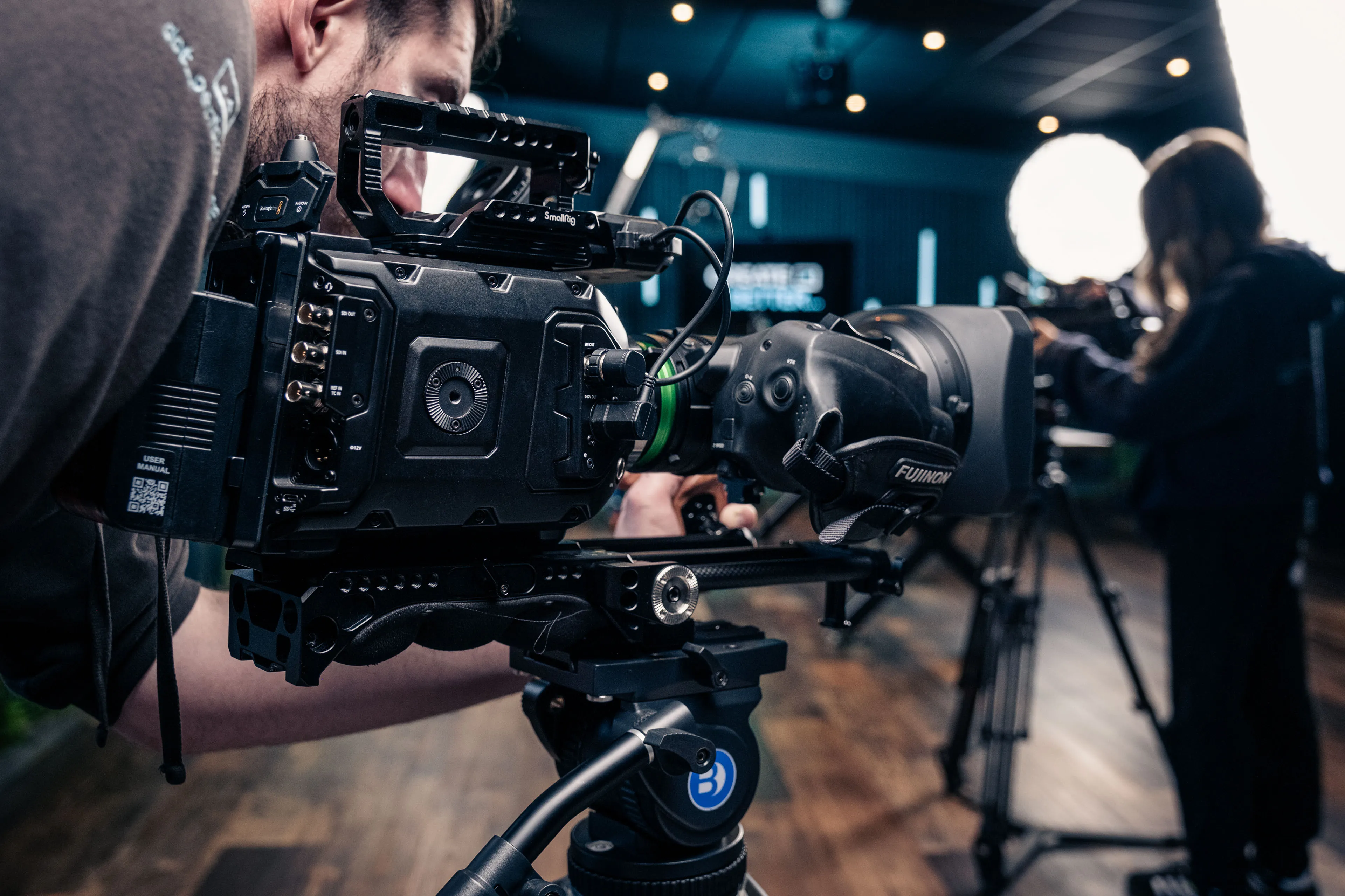 A cinematographer peers through a professional digital cinema camera mounted on a tripod in a dimly lit studio with bright background lights.