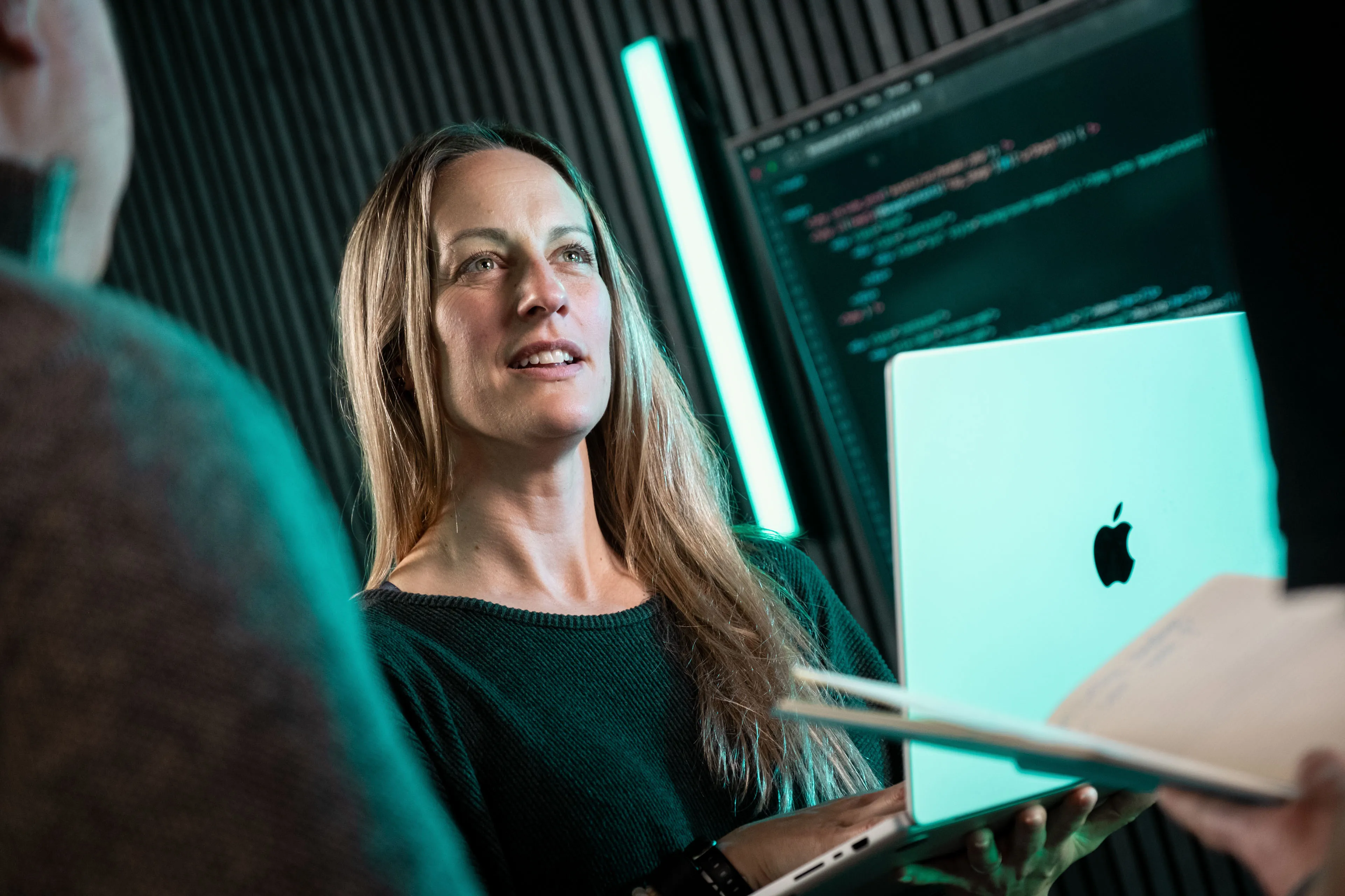 Rachel Fabrizio holding an Apple MacBook in conversation with a colleague at DotPerformance studios on the Isle of Man, with front-end code visible on a large screen behind her