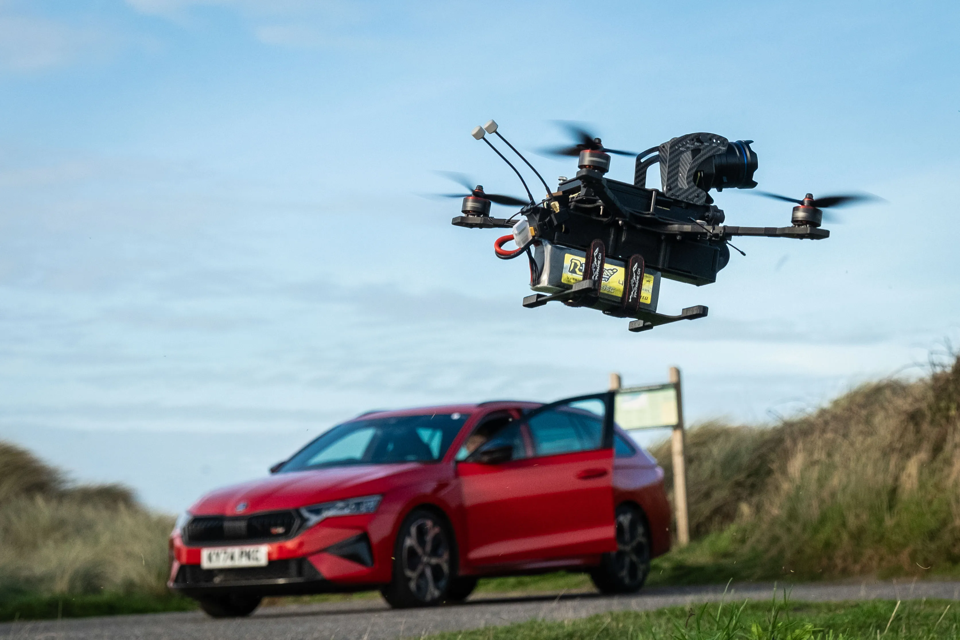 A drone hovers mid-air against a blue sky, with a red Skoda car parked on a rural road in the background. The scene conveys a tech-savvy and adventurous tone.