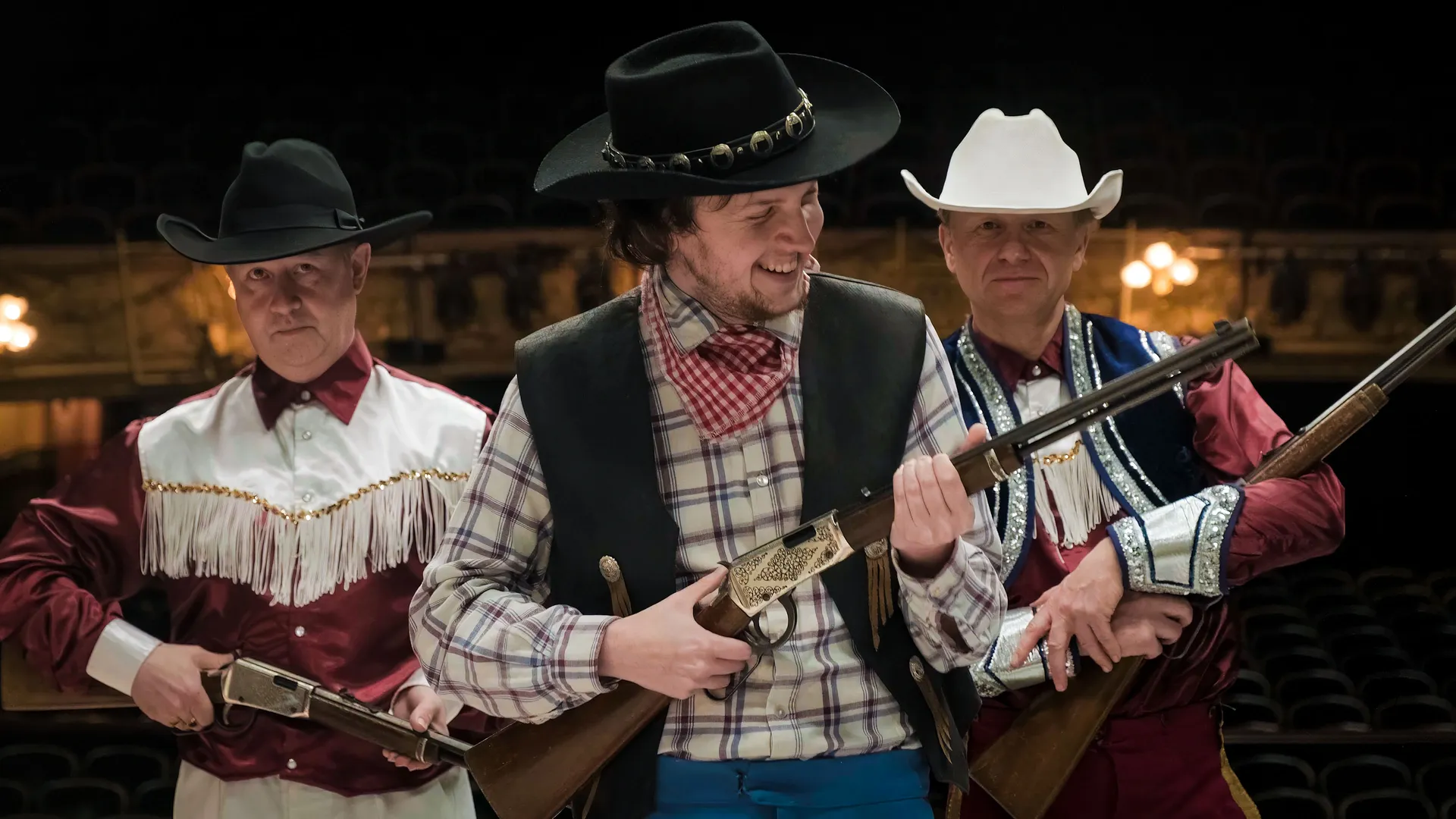 Three men dressed as cowboys brandish rifles on stage. The central figure in a black hat grins, exuding confidence. Theater setting, playful mood.