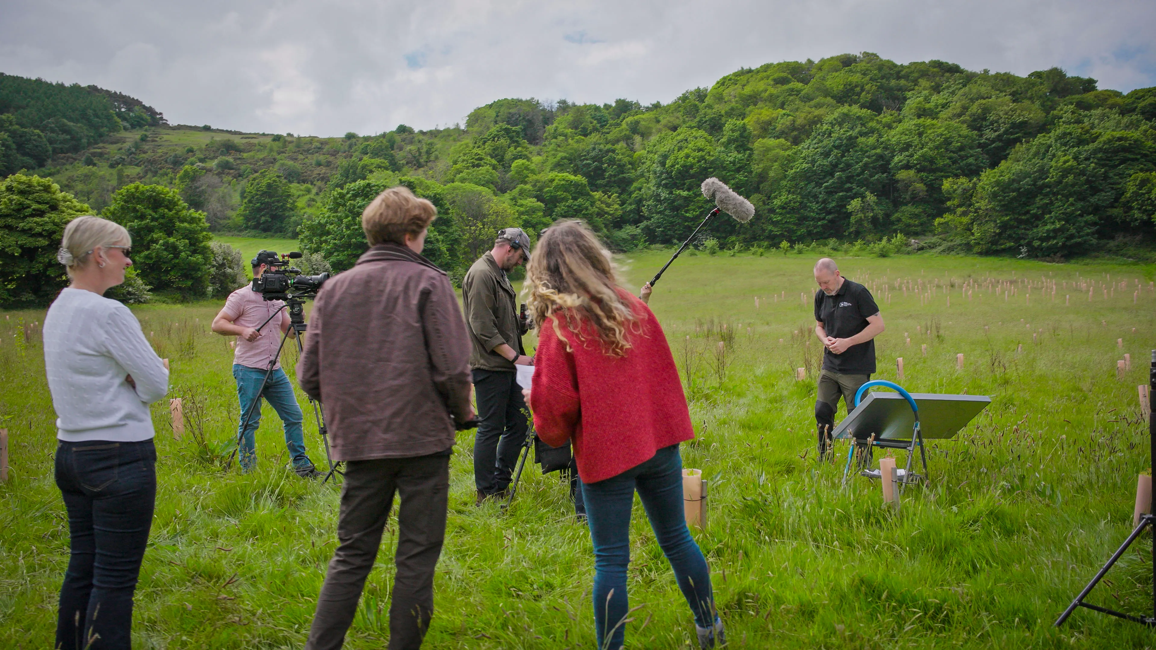 A film crew in a lush, green field with wooded hills in the background. People hold a camera and boom mic, capturing a person explaining something in the field.