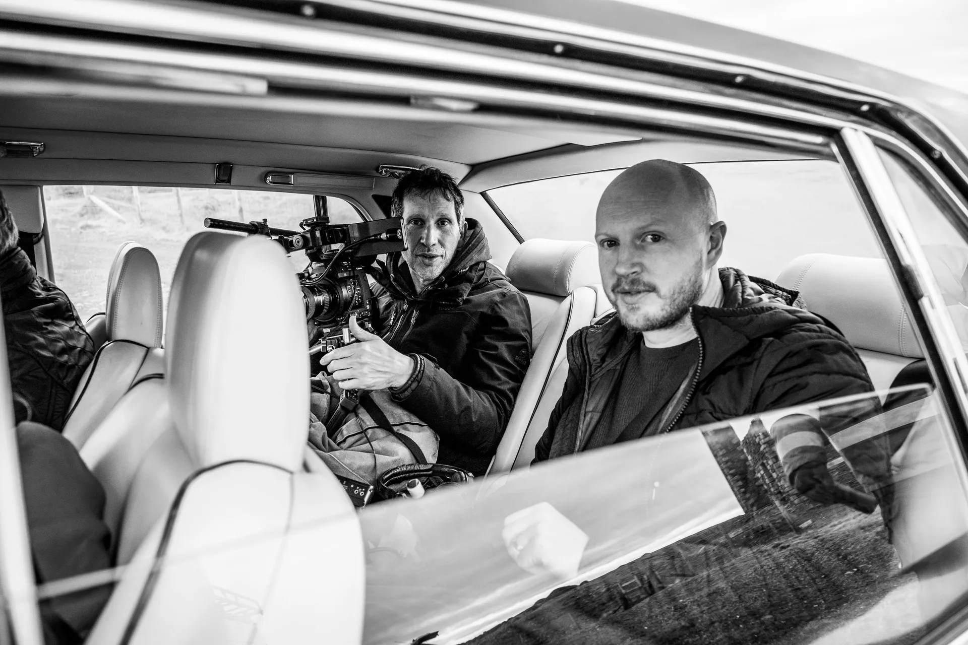 Two men sit in a parked car; one is operating a video camera, the other looks towards the camera, both in black jackets, creating a focused tone.