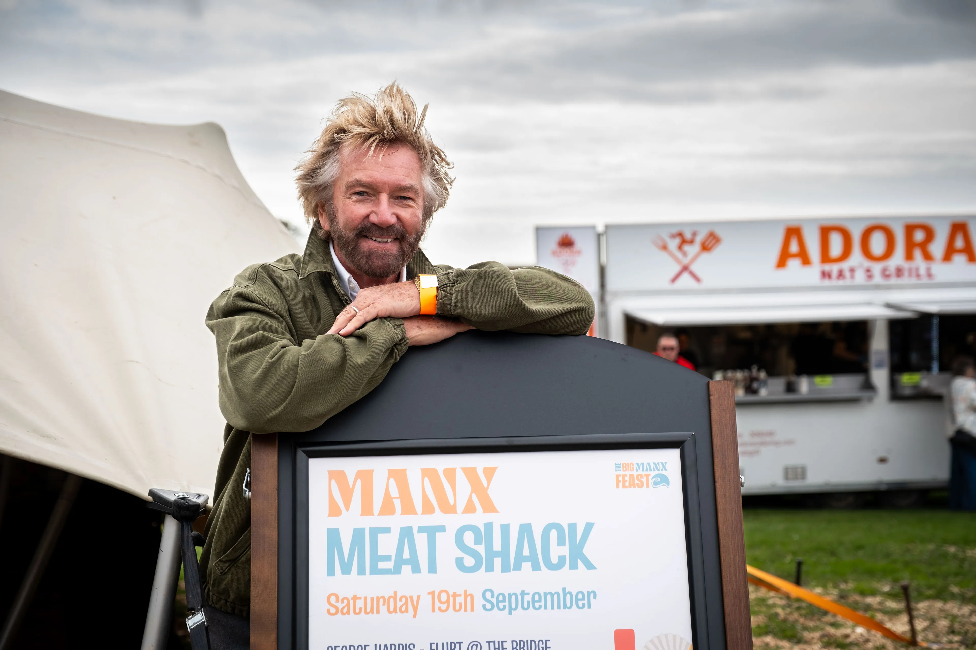 Noel Edmonds leaning on the Manx Meat Shack sign at the Big Manx Feast, with Adora Nat's Grill behind him