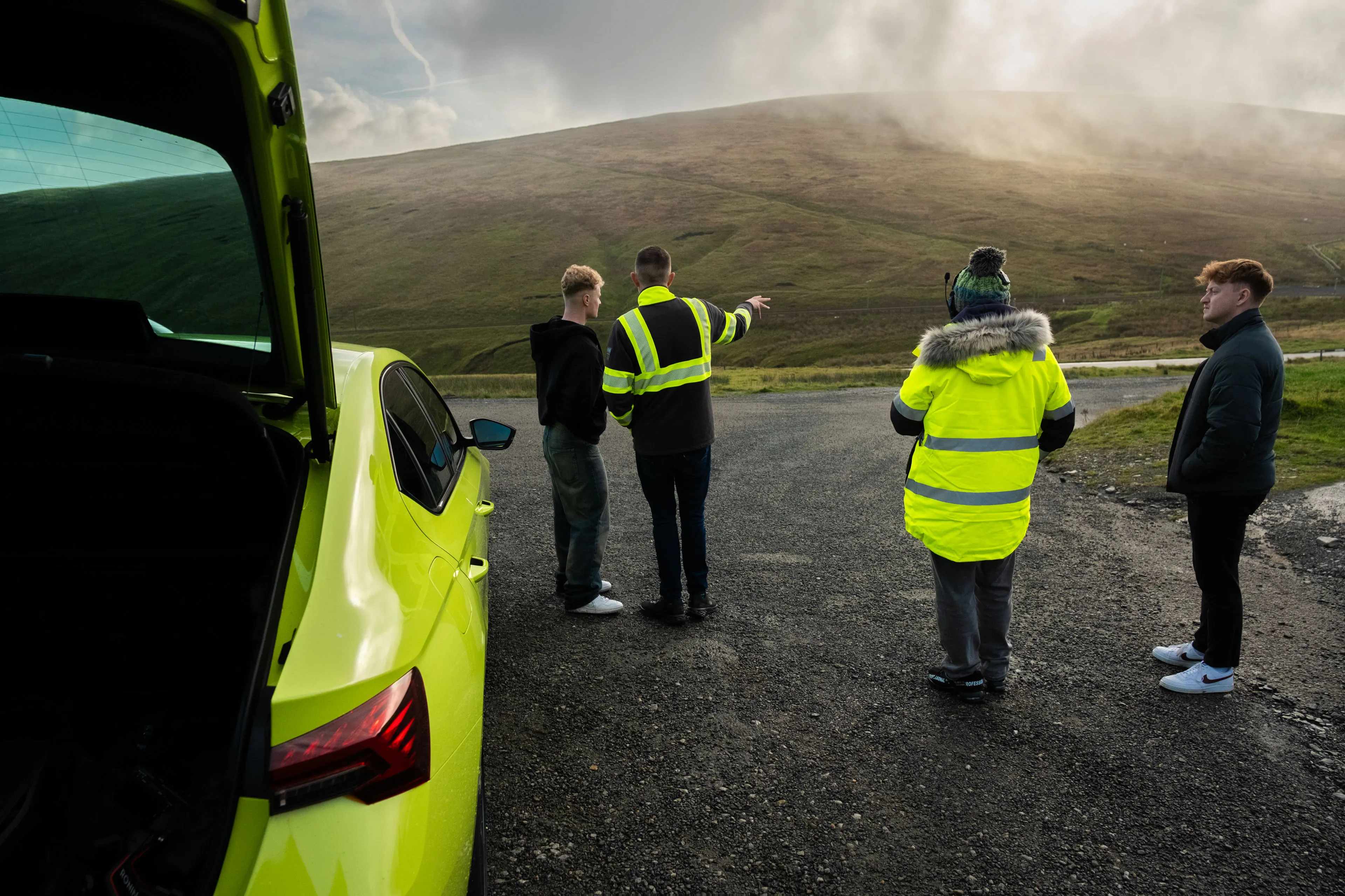 Four people, two in bright high visibility jackets, stand near a neon green Skoda car on a rural road. One person points toward a misty, rolling hill.