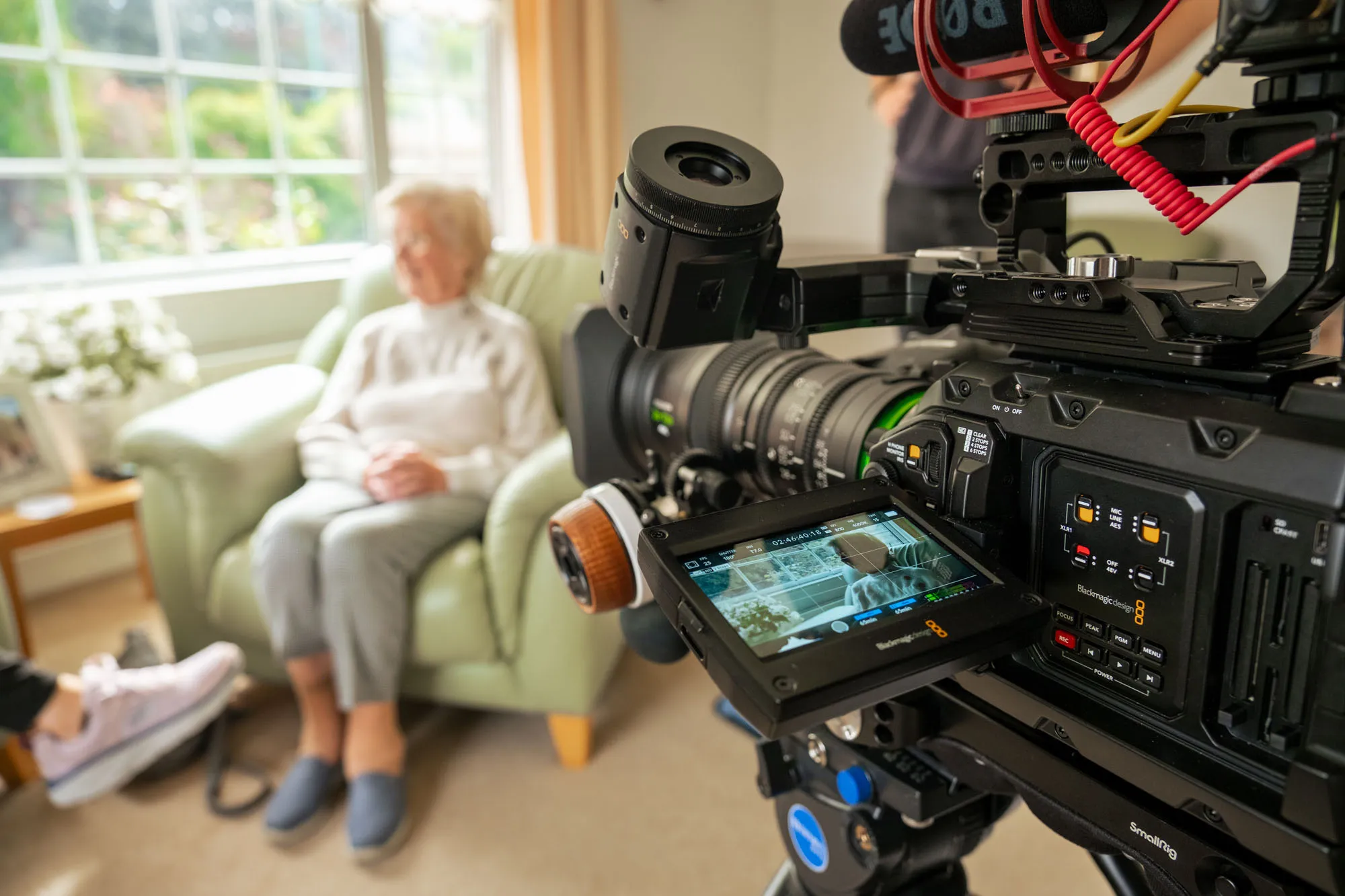 A professional video camera focuses on an elderly woman seated on a green armchair in a cozy, sunlit room, creating an atmosphere of calm and focus.