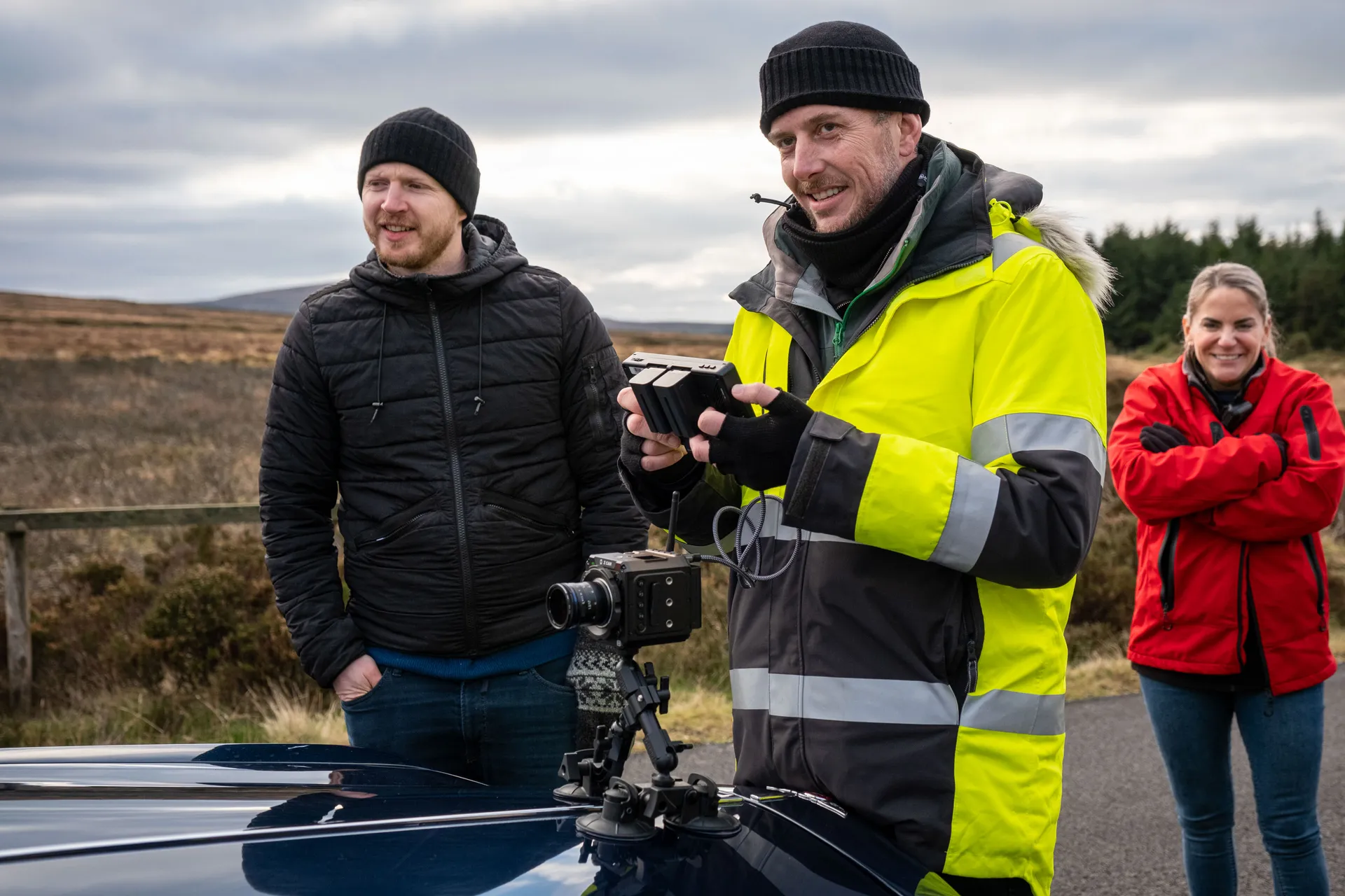 Three people stand outdoors on a cloudy day. One in a bright yellow coat operates a camera mounted on a vehicle. The mood is casual and focused.