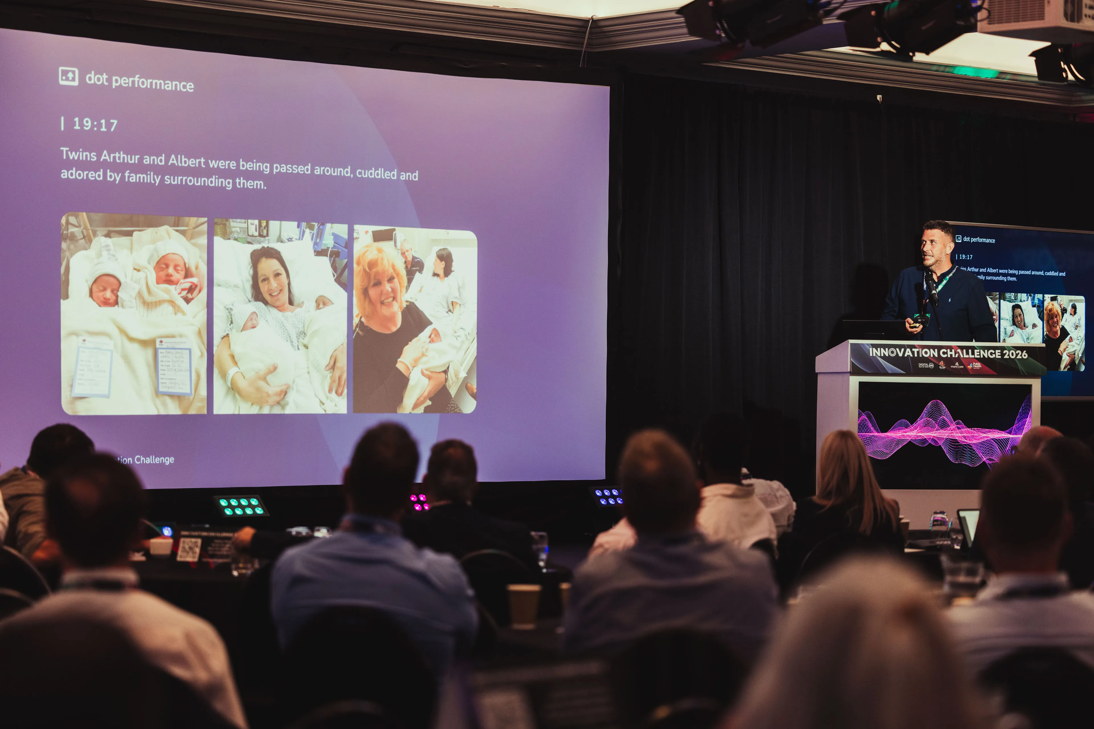 Andy Orton presenting beside a dot performance slide showing twin newborns being cuddled by family in hospital