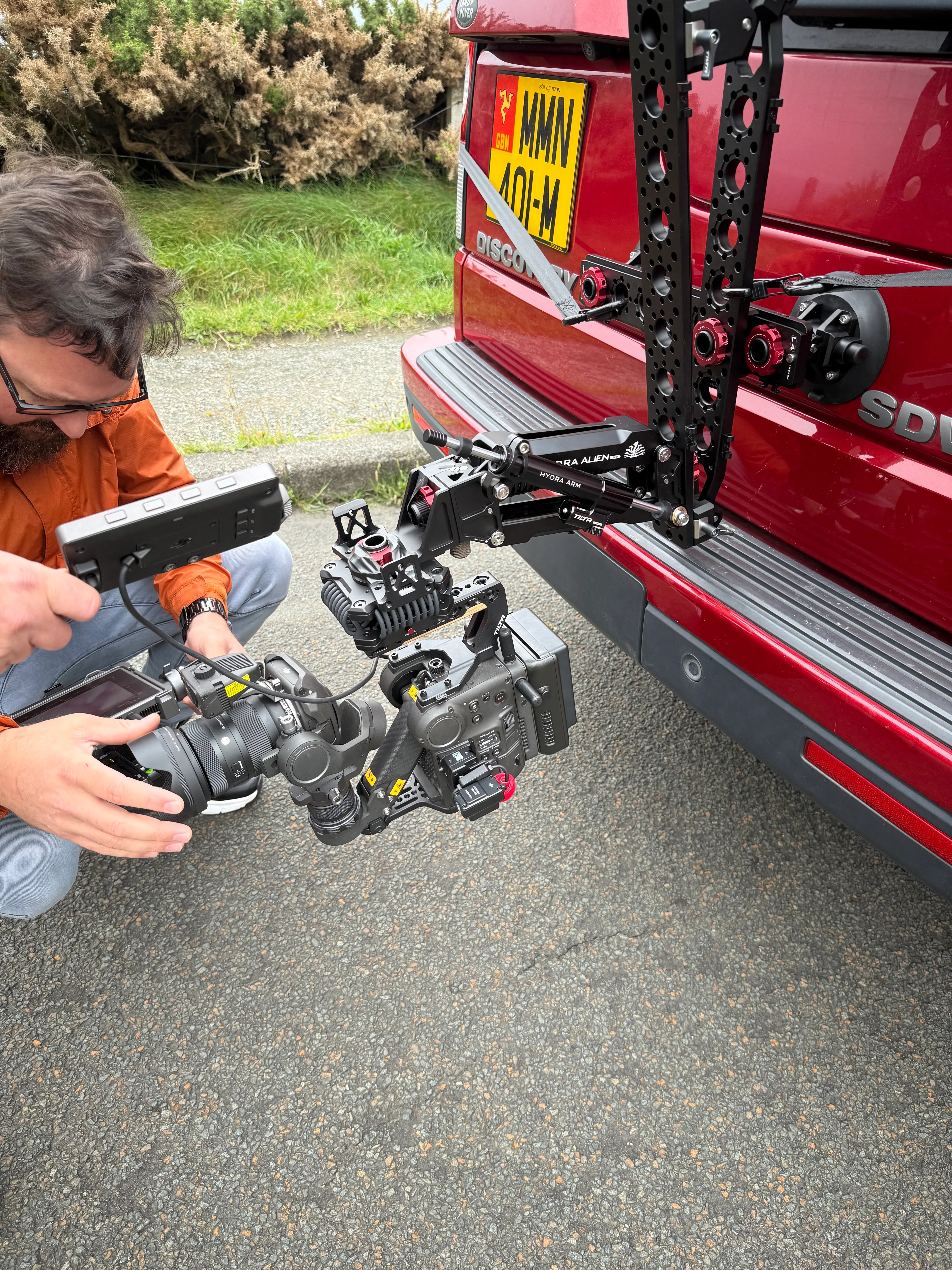 Nico van Loggerenberg checking a DJI Ronin 4D mounted on a Tilta Hydra Alien arm at the rear of a red vehicle on the Isle of Man during pre-shoot setup