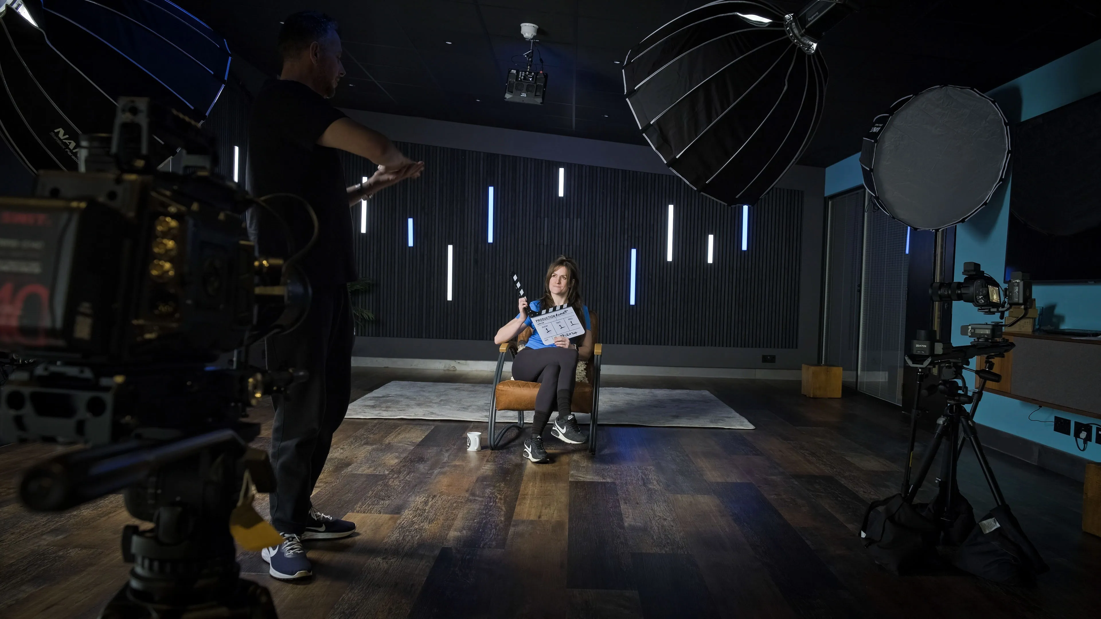 Film set with a cameraman directing a seated woman holding a clapperboard. Studio lights and cameras surround them, creating a focused, professional atmosphere.