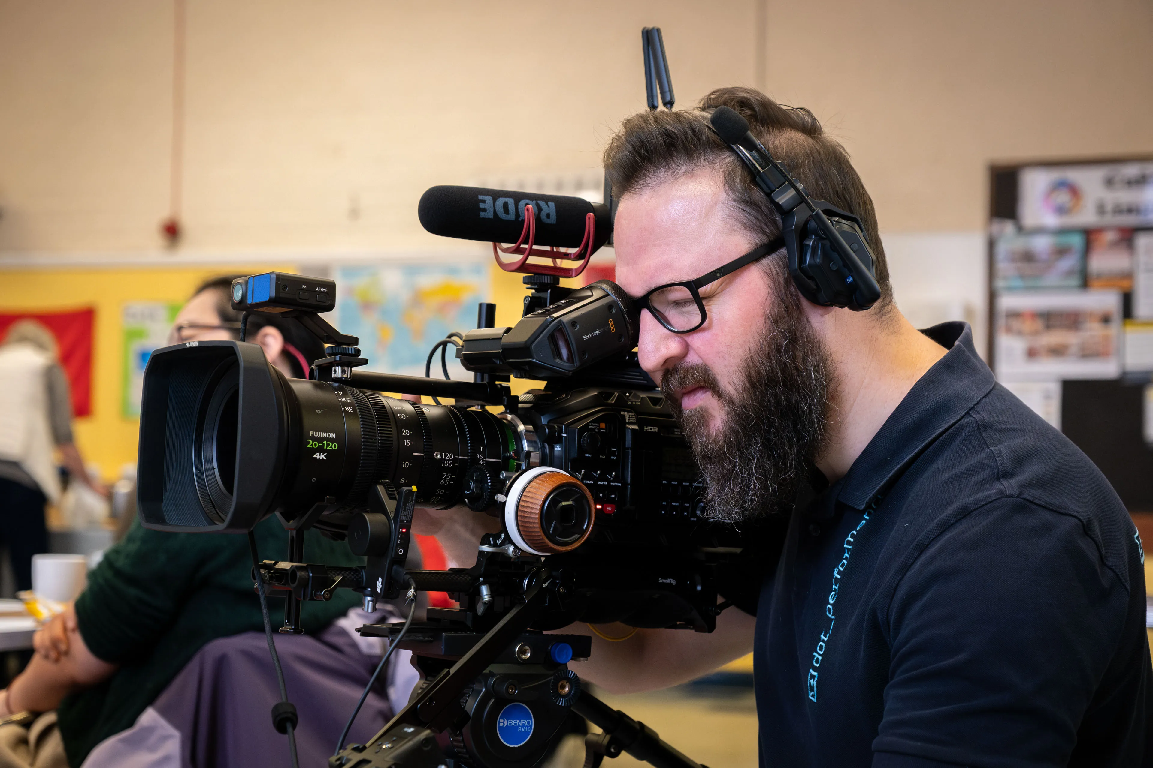 A bearded cameraman in glasses and headphones operates a professional video camera indoors, focusing intently. Background features colourful maps and posters.