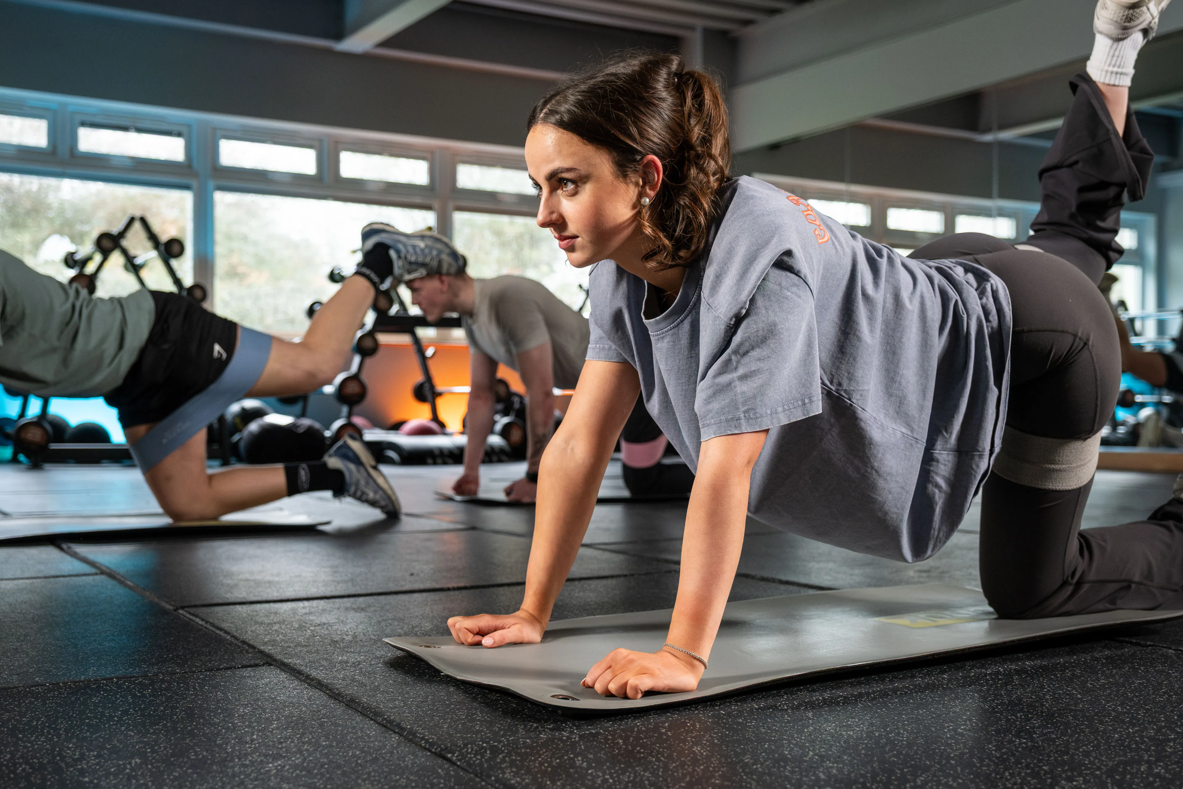 A focused woman in a gym class performs a leg raise on all fours, wearing a blue shirt and black leggings. Other participants exercise in the background.