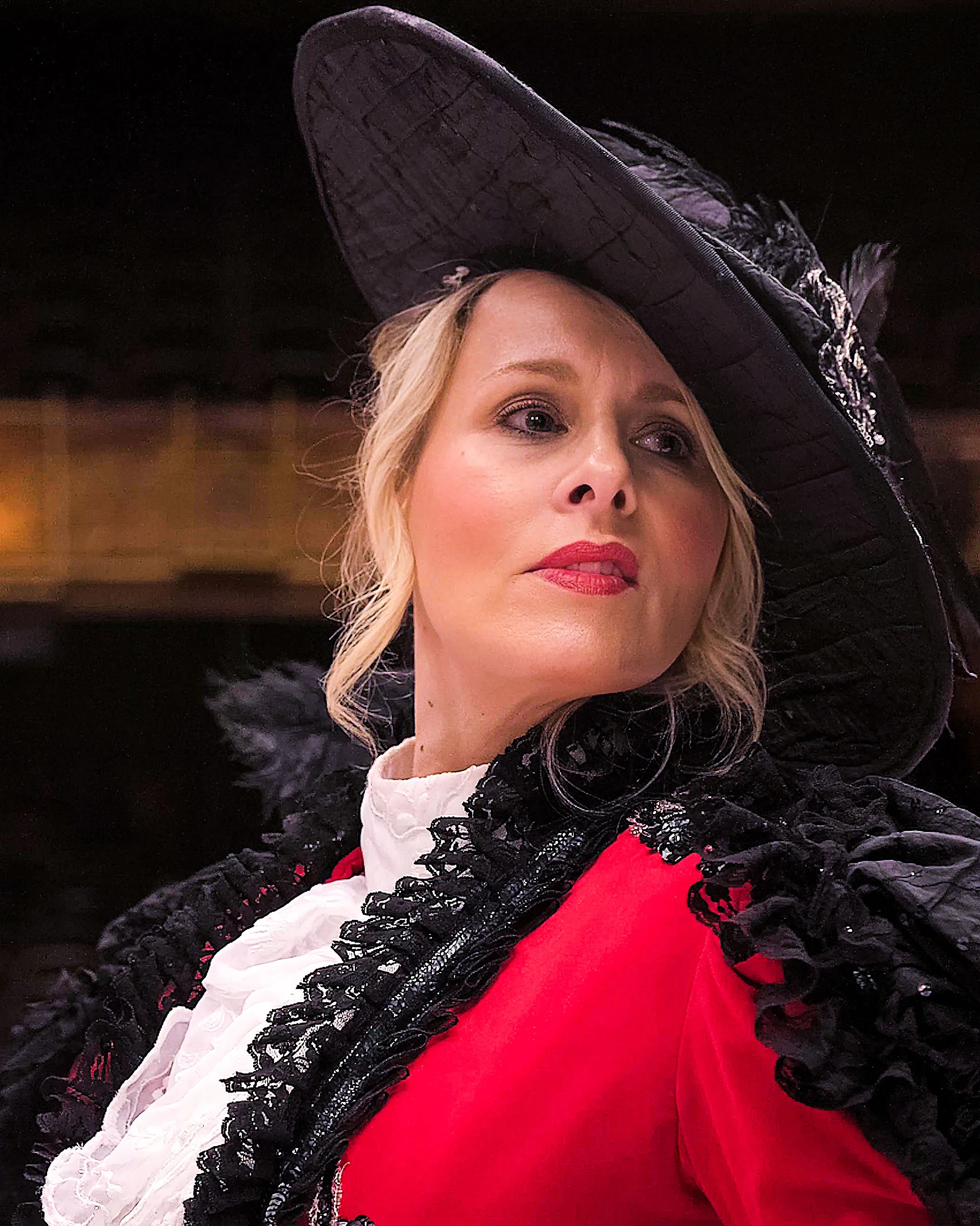 A woman dressed in an elaborate historical costume gazes confidently to the side. She wears a large black hat with feathers, a red coat, and a lace-trimmed blouse.