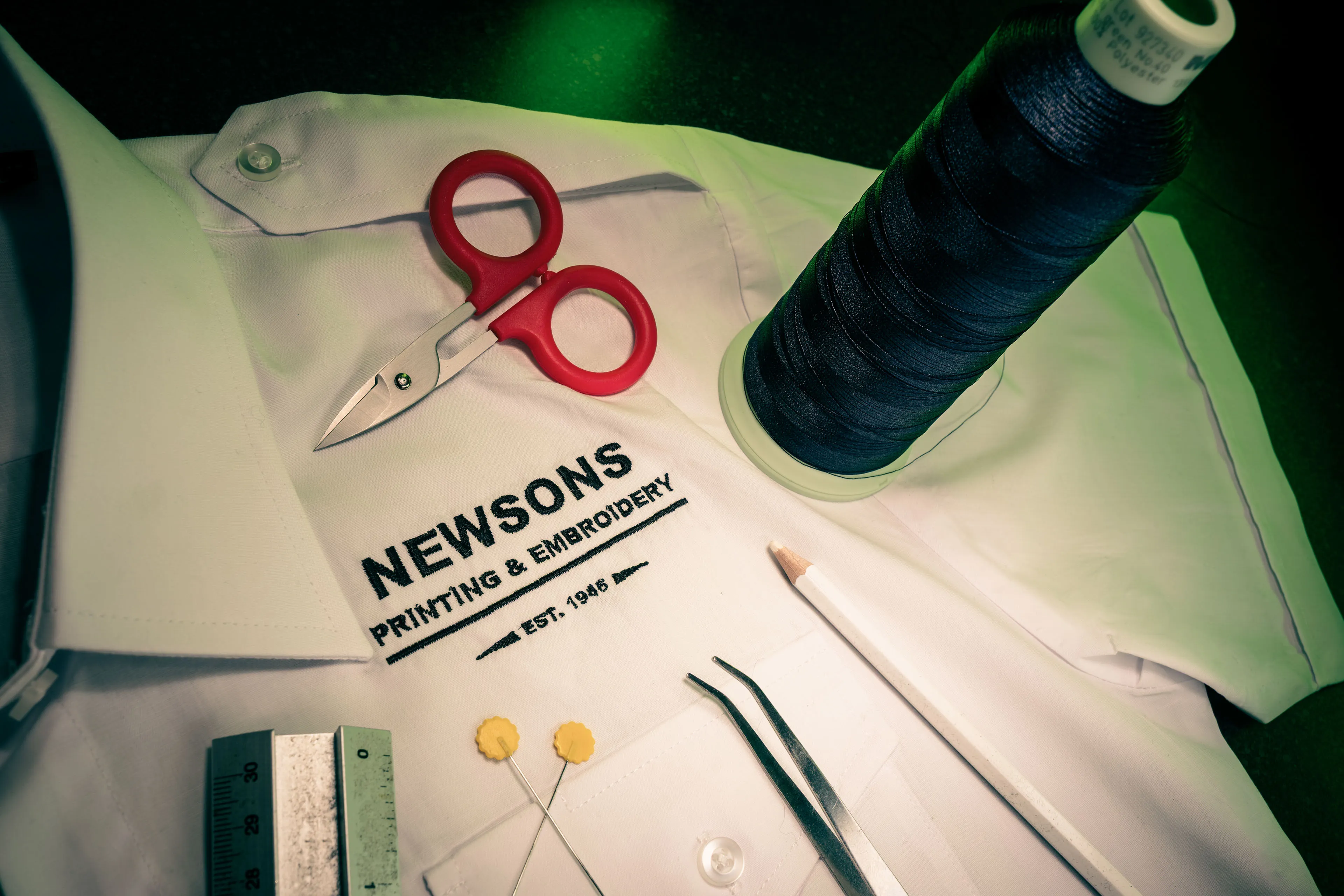 A white shirt with "Newsons Printing & Embroidery" logo is surrounded by red-handled scissors, a spool of blue thread, a ruler, tweezers, and pins.
