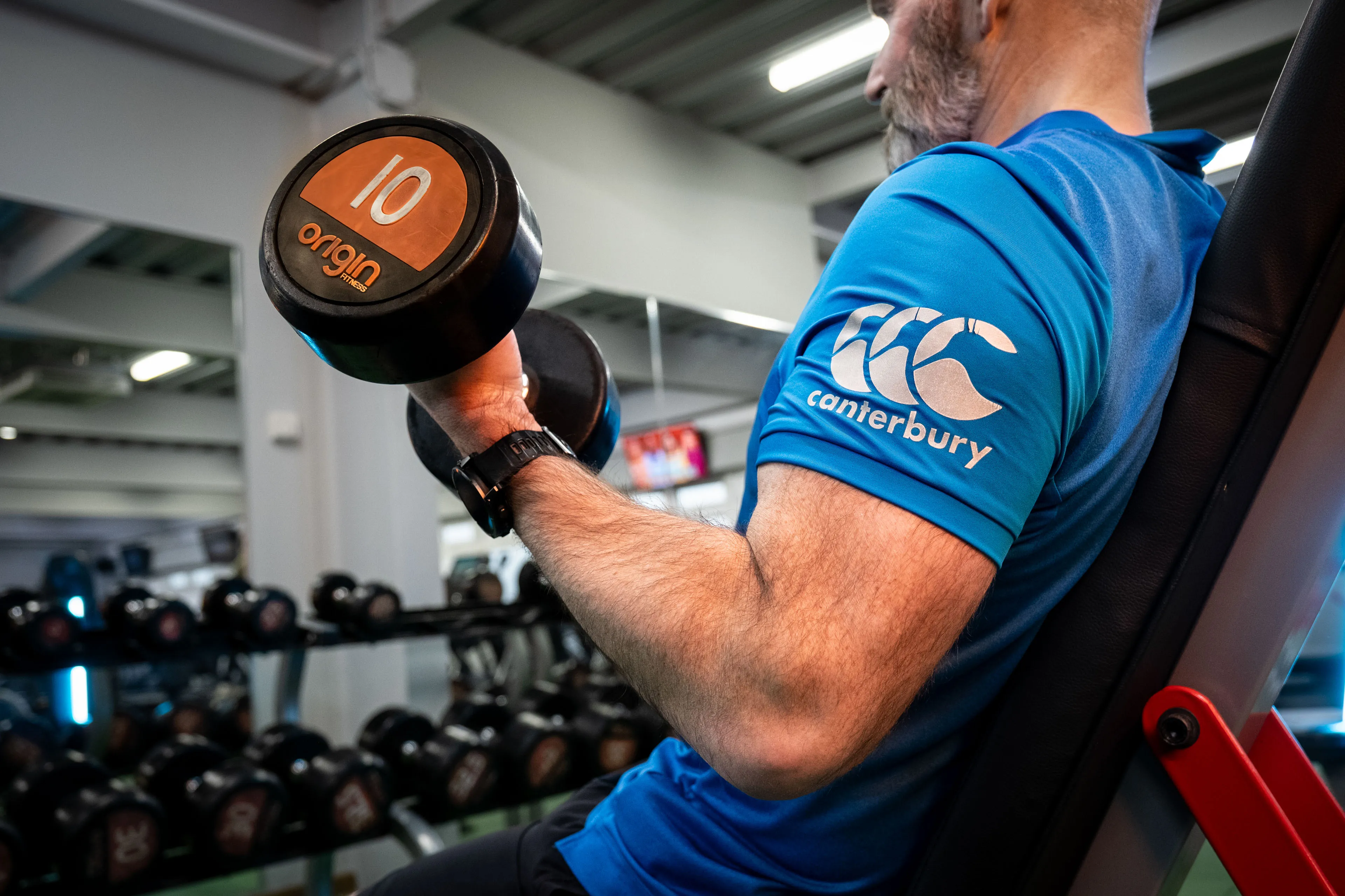 Man lifting a 10 kg dumbbell at the gym, wearing a blue shirt with a Canterbury logo. Background shows rows of dumbbells. Focus on strength and fitness.